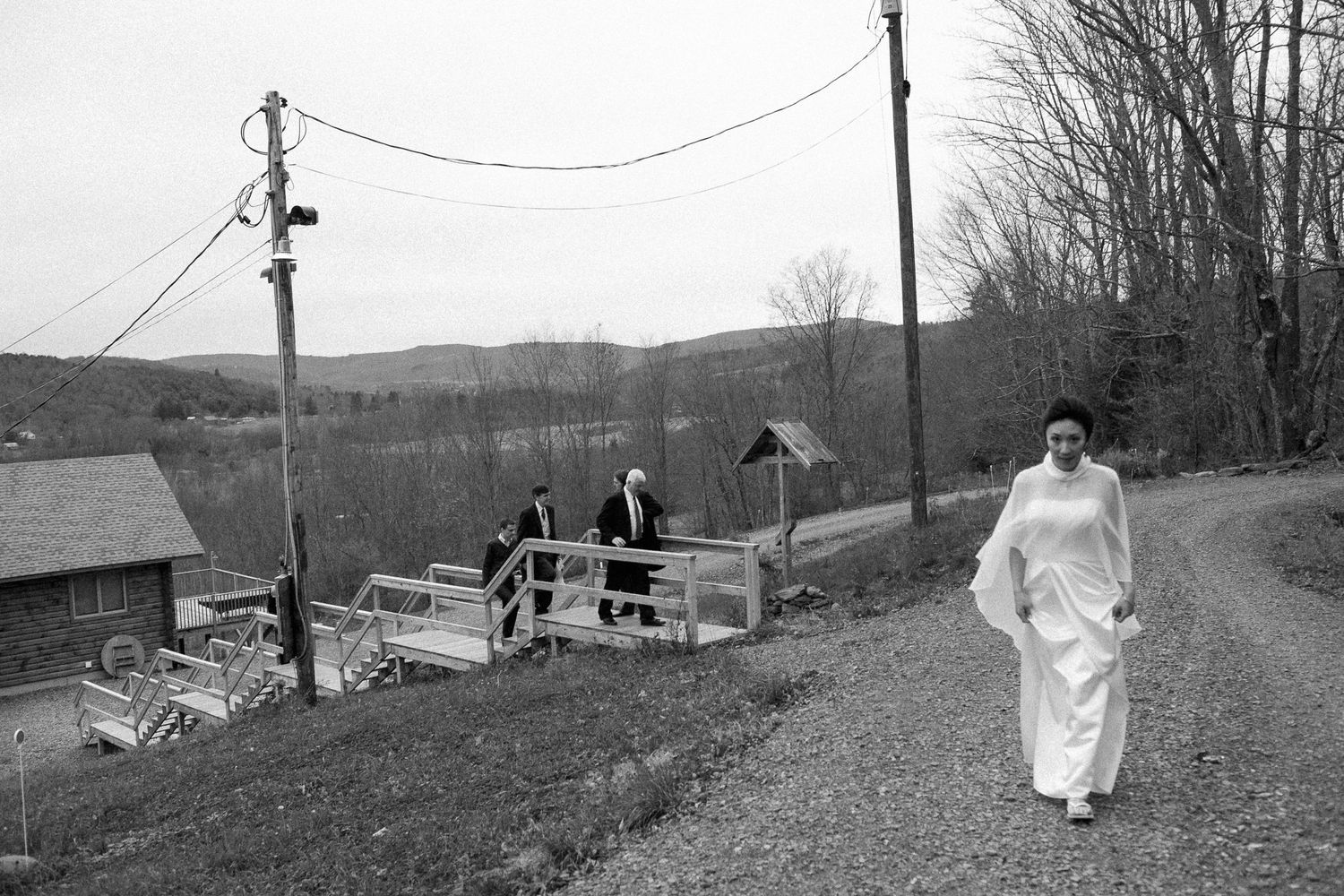 Black and white photo of a woman in white dress walking rural gravel road with others near a wooden fence.