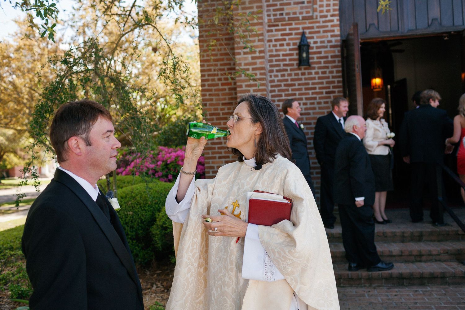 Priest in white vestments drinking from a bottle while speaking with a groom outside a church wedding ceremony.
