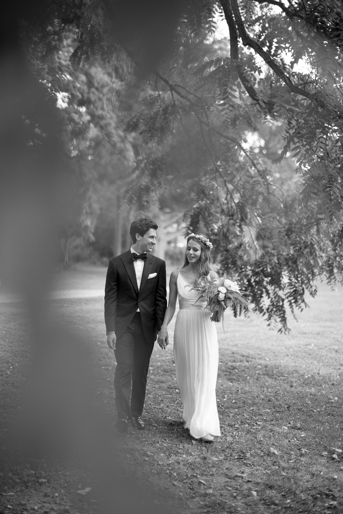 Black and white portrait of wedding couple walking under trees in a park.