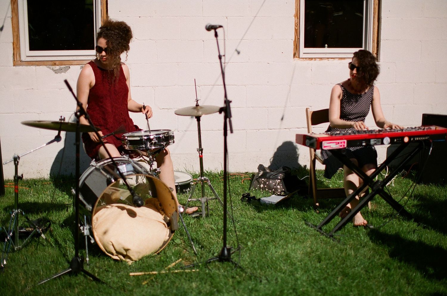 Two musicians perform outdoors, one playing drums and one on keyboard, on a sunny day against a white building.
