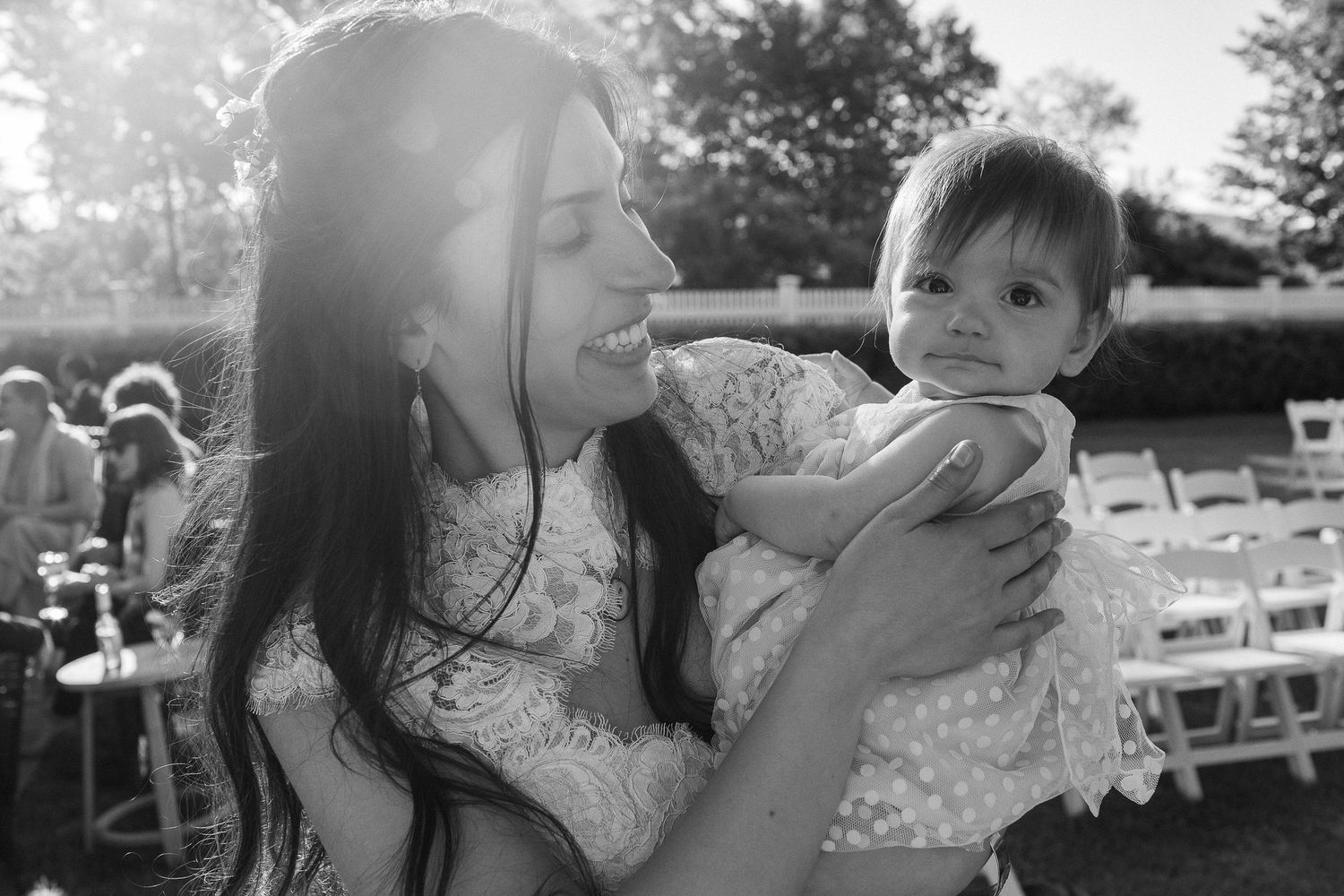 Black and white photo of a smiling woman holding a cute baby at an outdoor wedding reception.