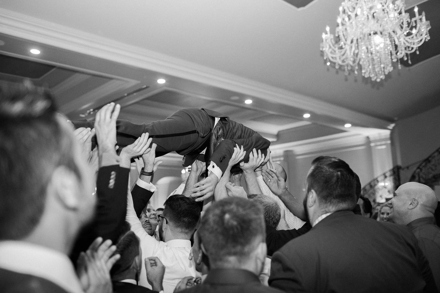Black and white photo of wedding guests lifting groom above crowd in festive celebration inside elegant ballroom.