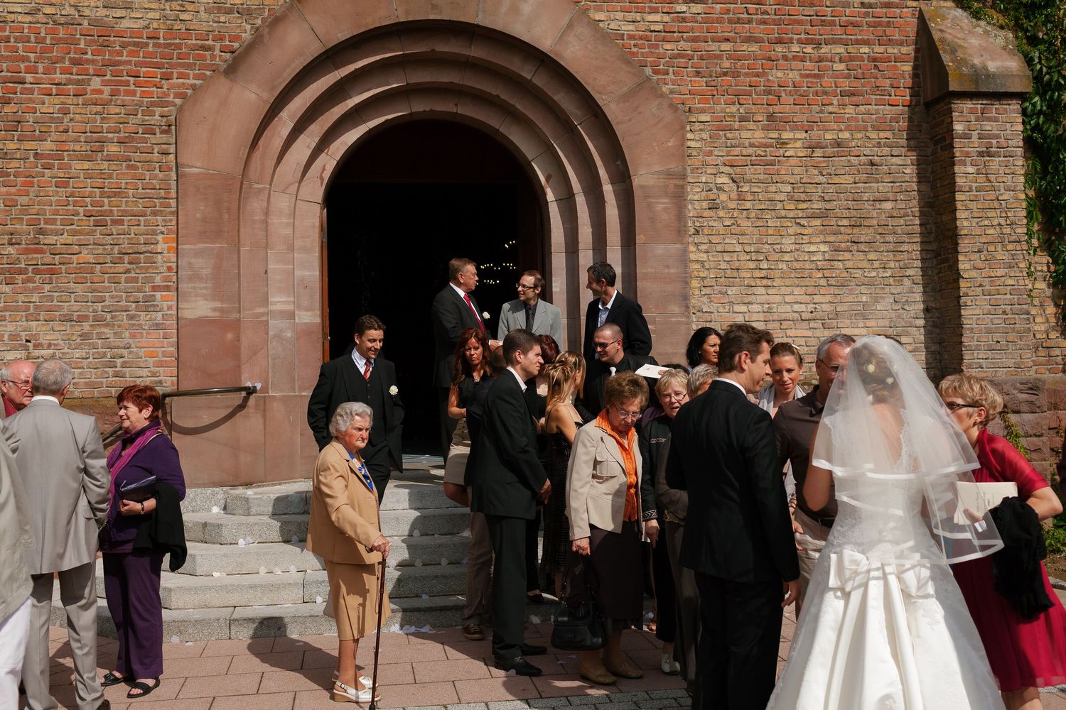 Wedding guests gather outside a brick church entrance after a ceremony, with the bride in a white gown visible on the right.