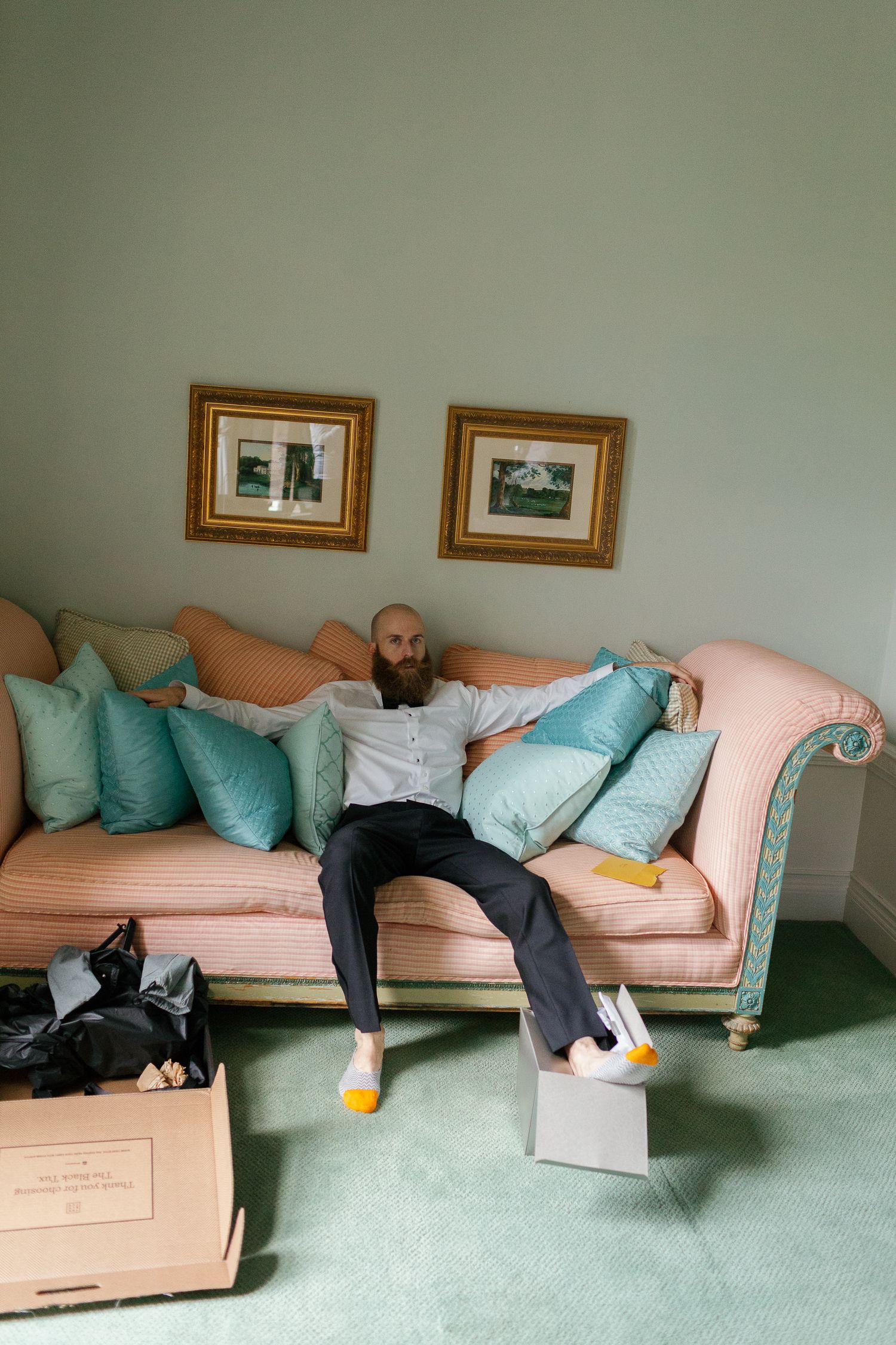 Bearded man relaxing on a pink sofa with teal cushions, wearing novelty socks, surrounded by moving boxes.
