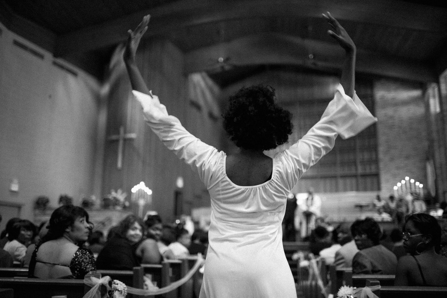 Black and white photo of a woman in white dress raising arms in praise inside a church congregation.