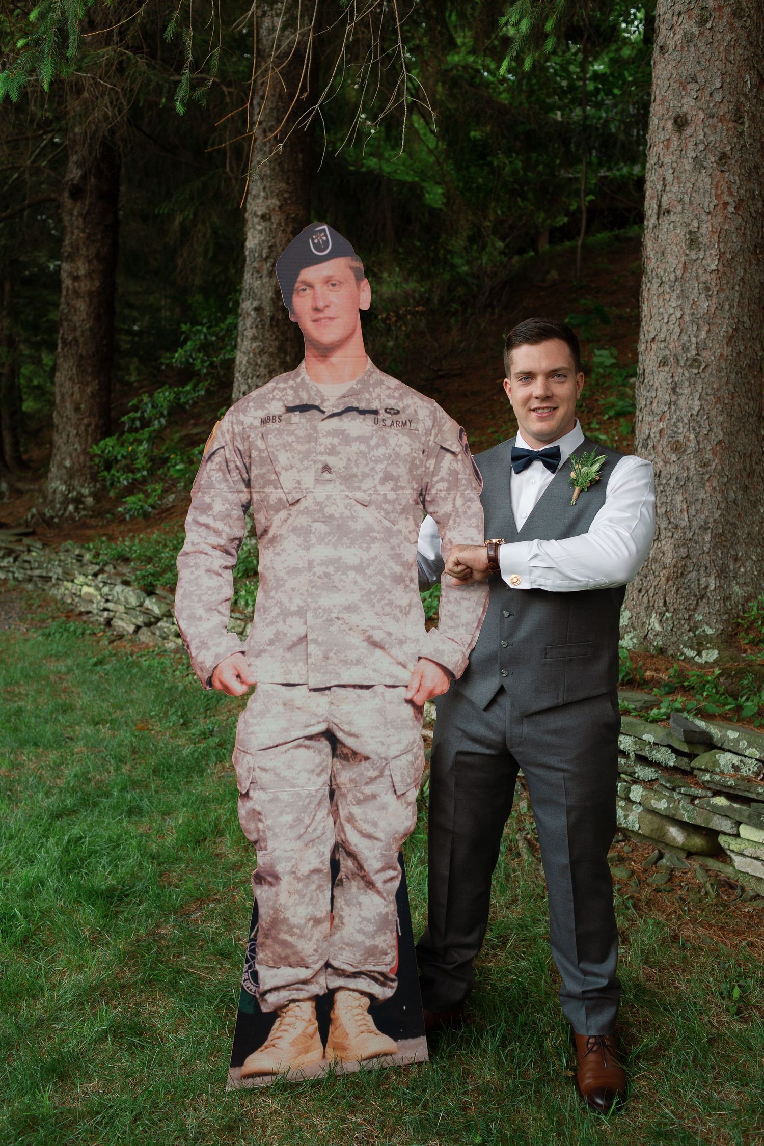 Young man in formal suit poses with a military soldier cardboard cutout outdoors near trees.