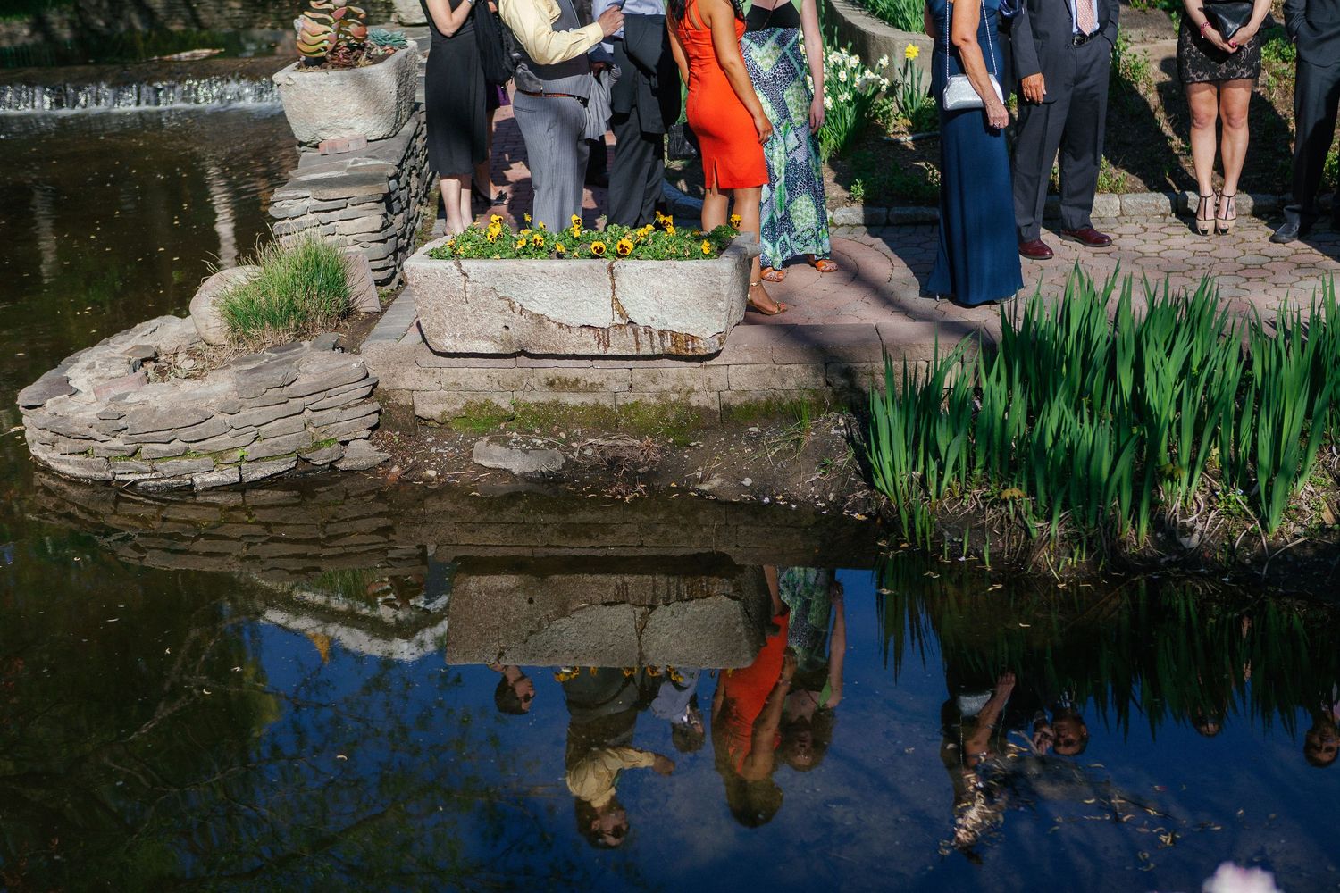 People standing by a reflective garden pond with lush greenery, their colorful reflections mirrored in the still water.
