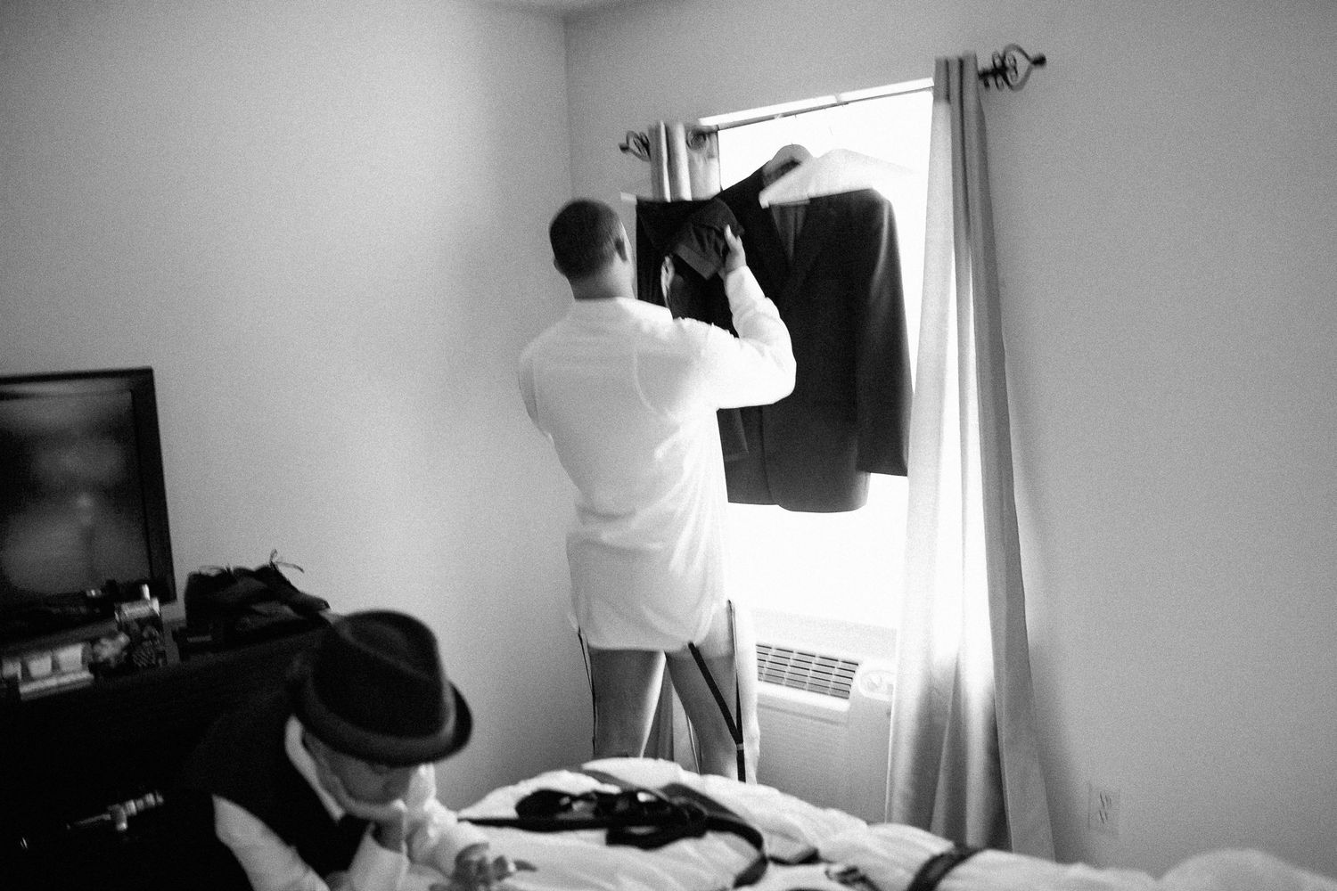 Man in white robe hanging dark suit jacket near window in bedroom, black and white wedding preparation photo.
