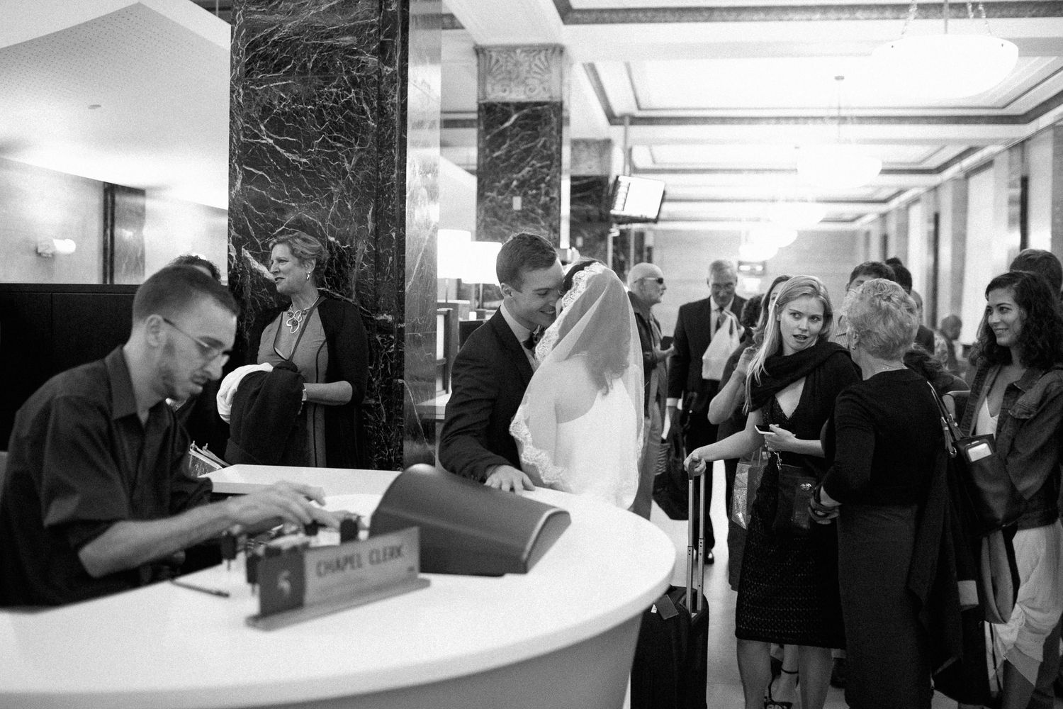 Black and white photo of a wedding couple checking in at a hotel reception desk surrounded by guests.
