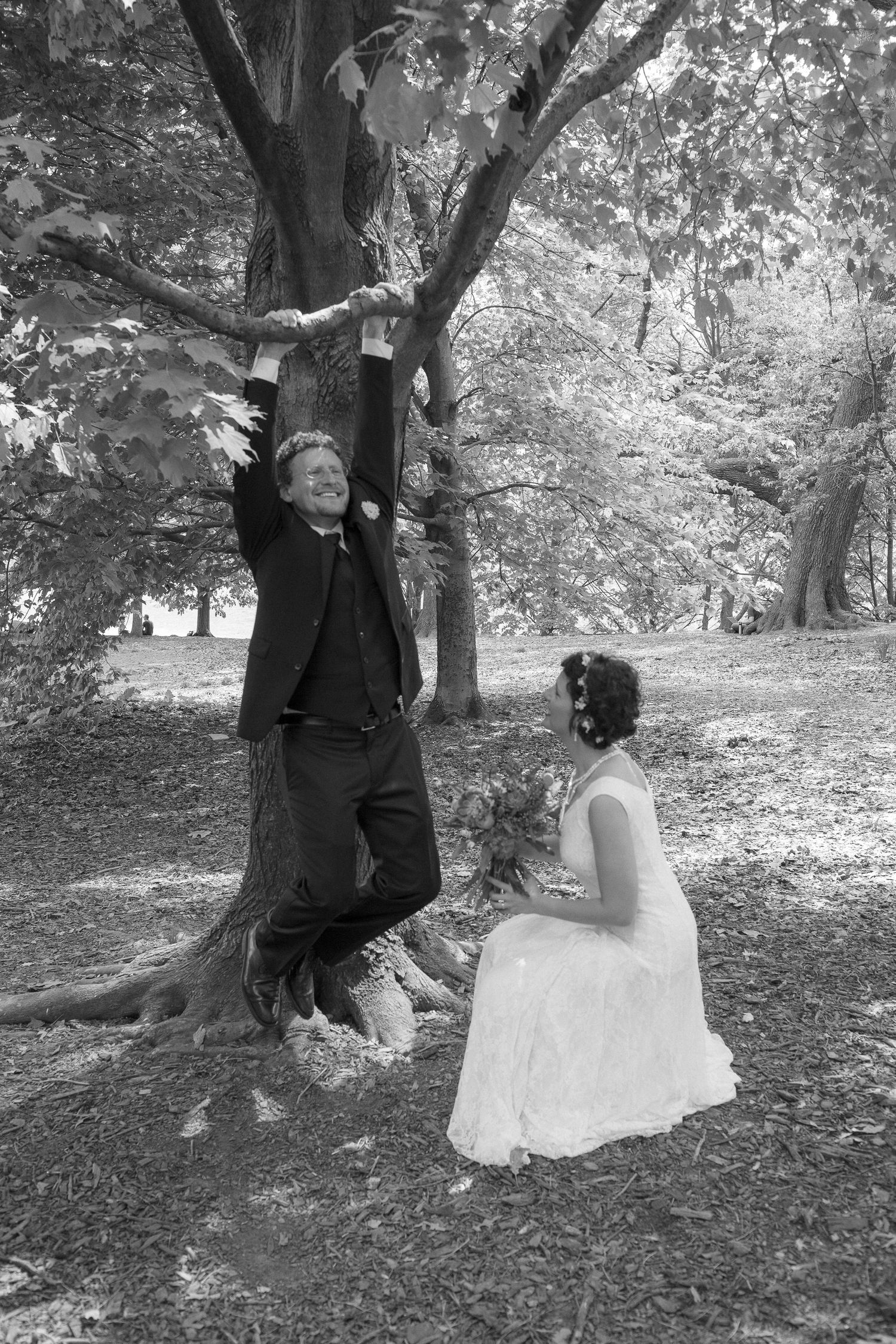 Black and white photo of bride and groom posing under a large tree in a wooded park setting.