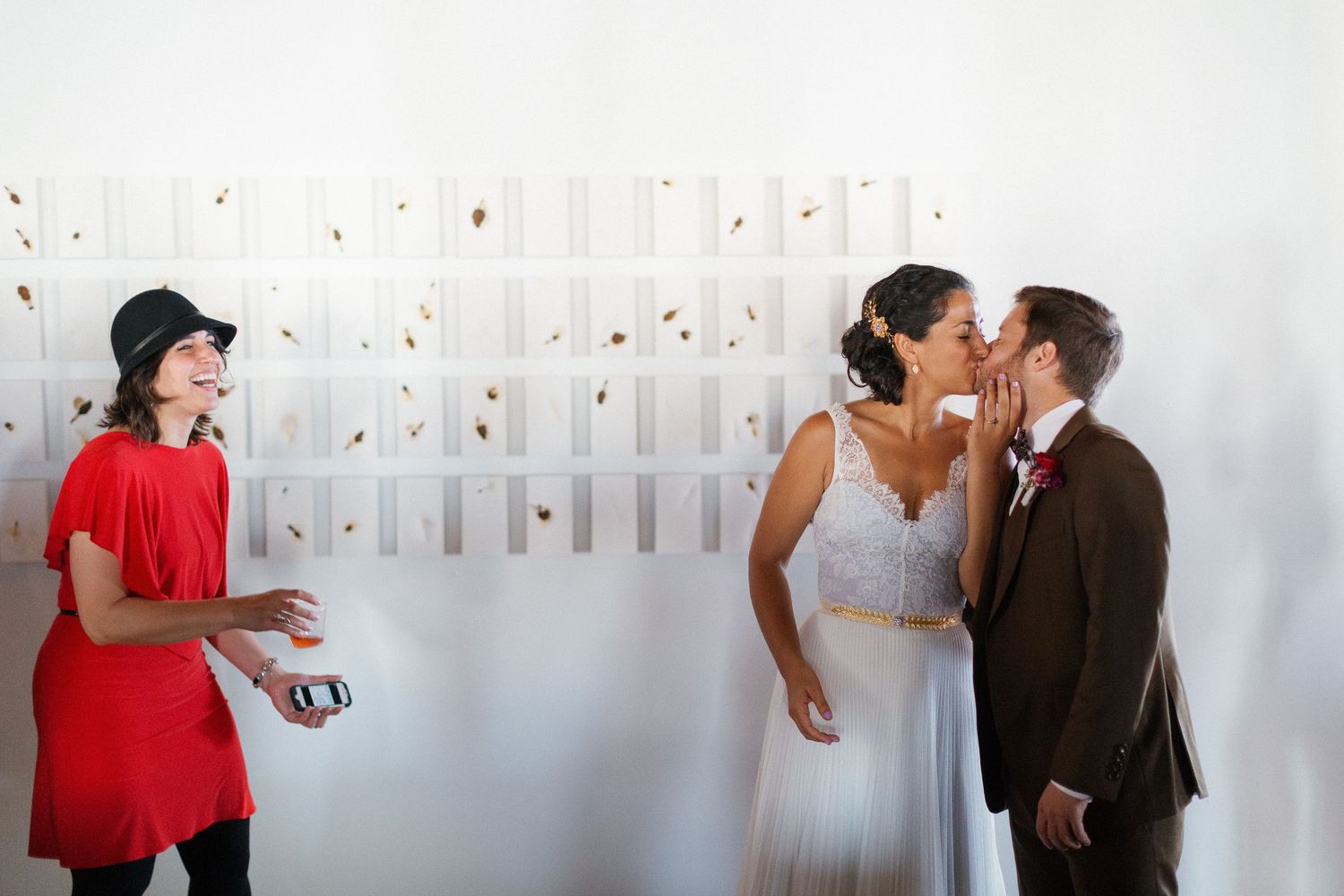 Bride and groom share a kiss at their wedding reception while a guest in a red outfit looks on.