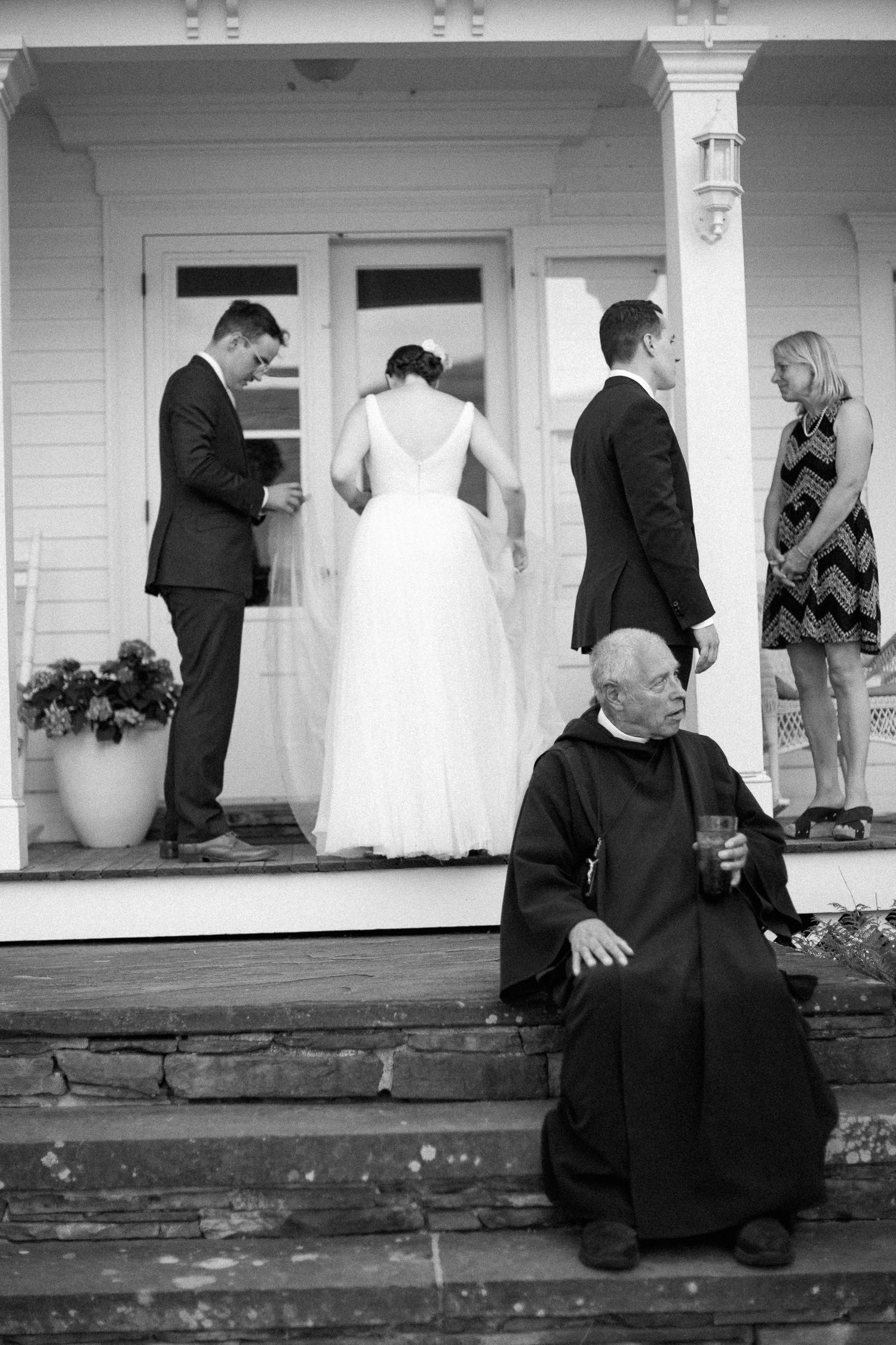 Black and white wedding photo of bride and groom on porch steps with guests, elderly man seated in foreground holding a glass.