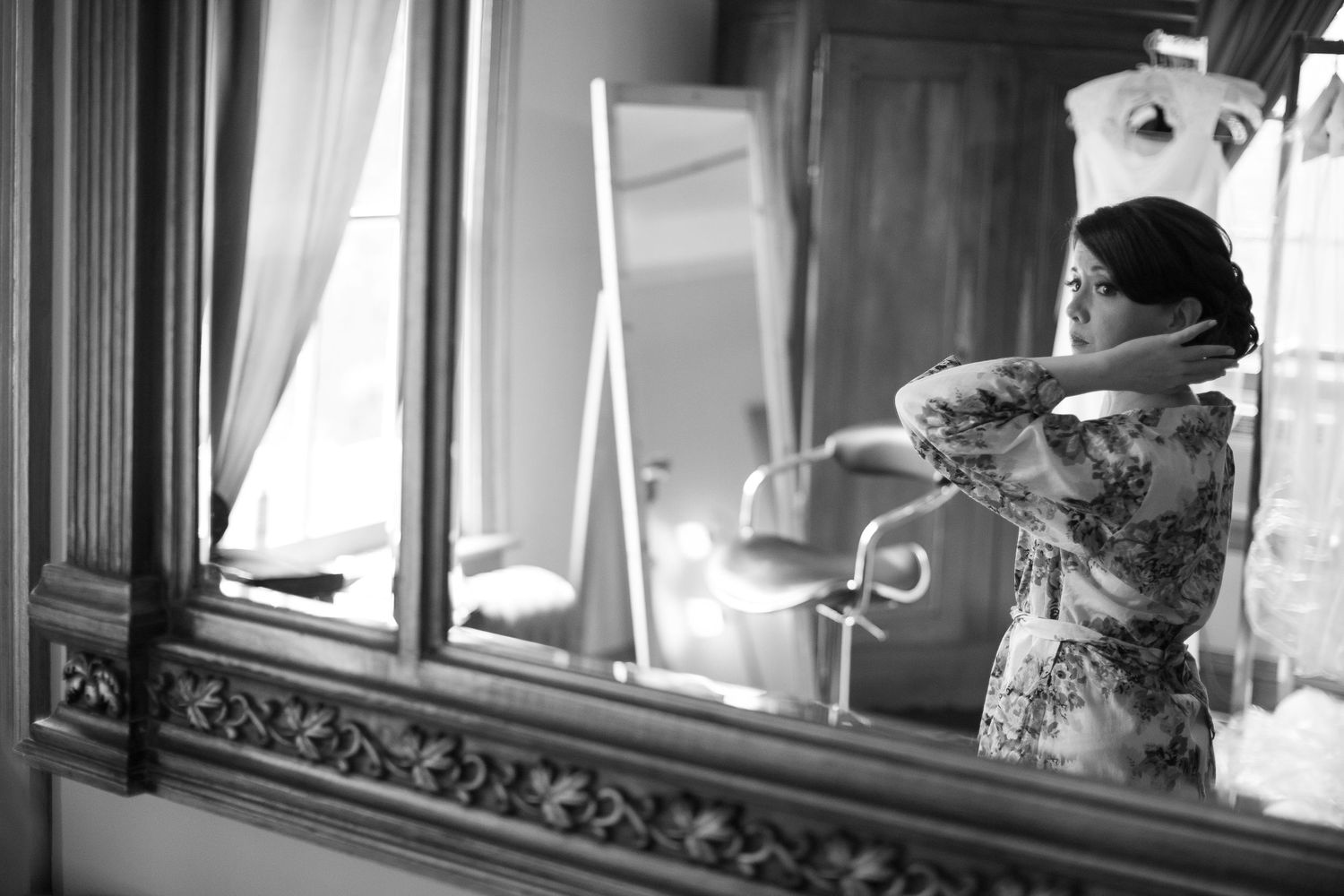 Black and white photo of a woman fixing her hair in front of an ornate mirror in an elegant room.