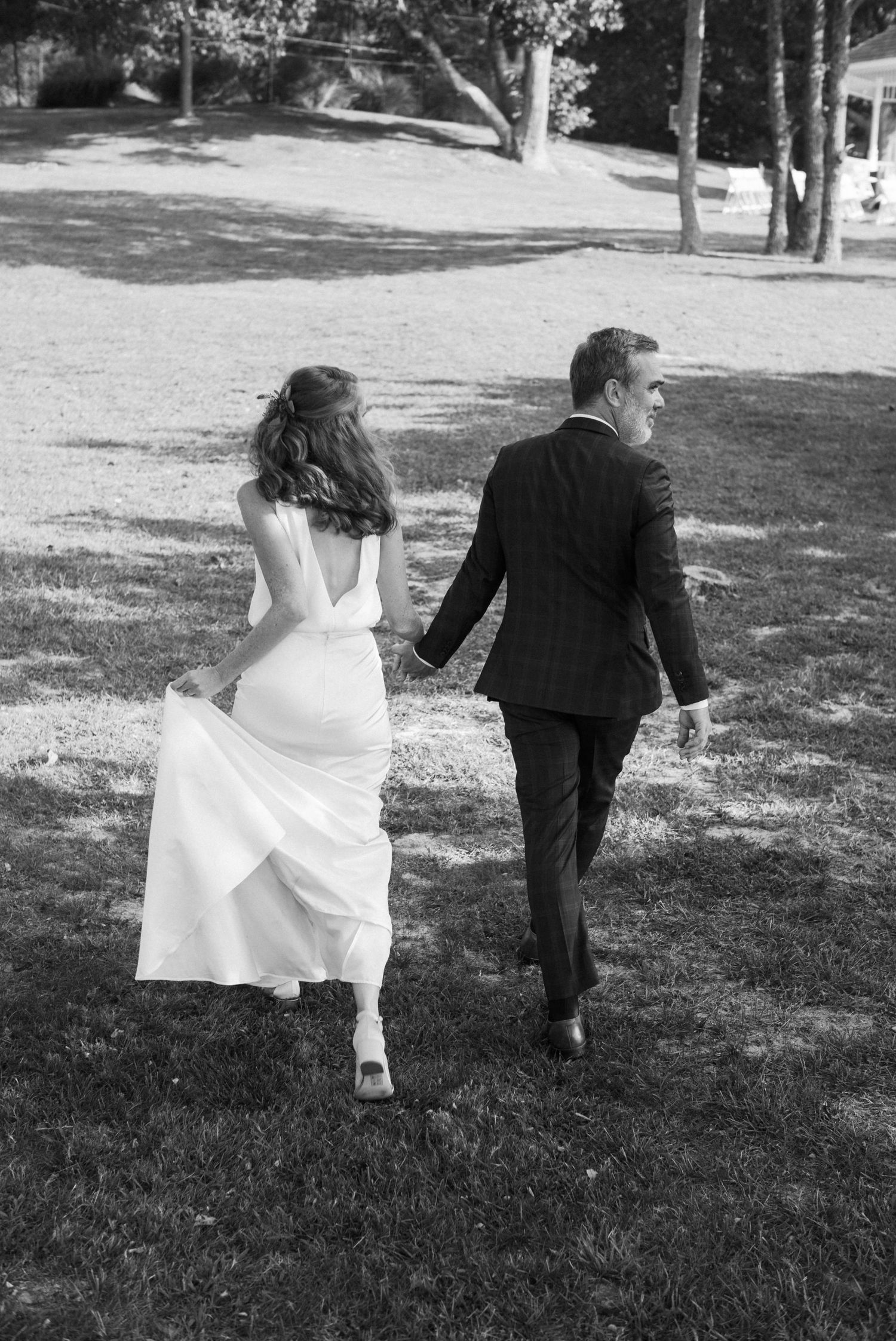 A black and white wedding photo showing a couple walking hand in hand across a grassy lawn on their special day.