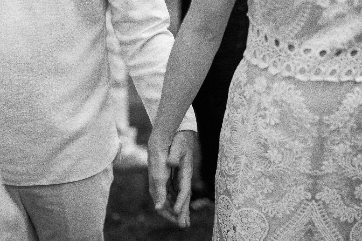 Black and white photo of a couple holding hands, bride in lace wedding dress beside groom in white shirt.