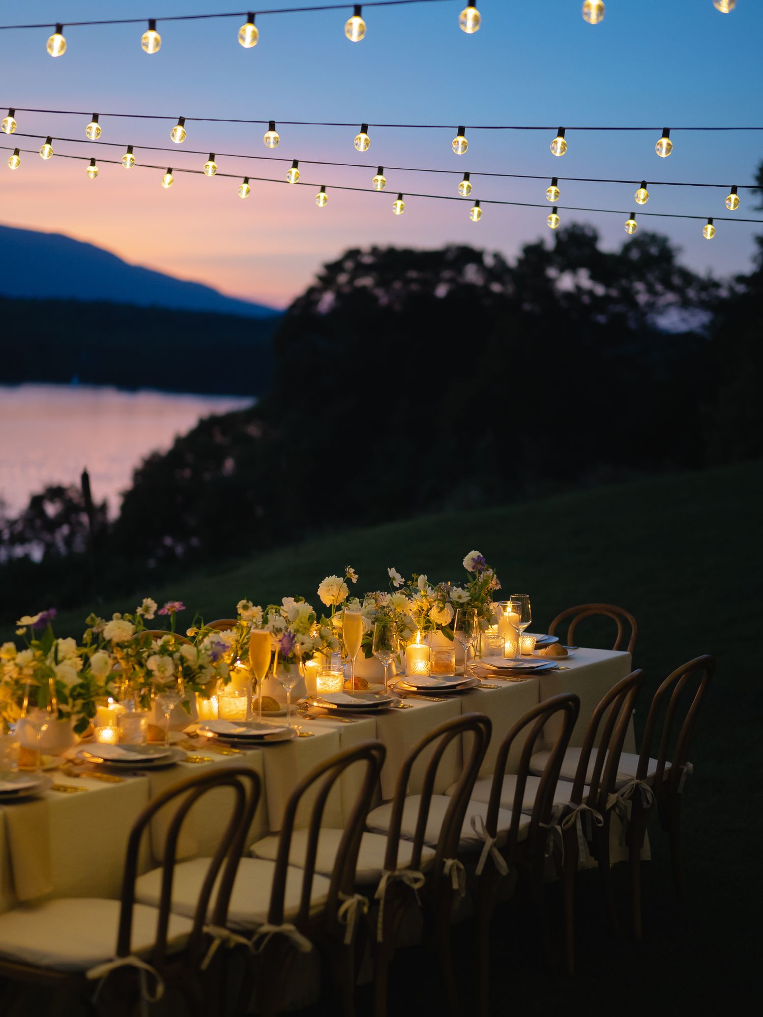 Romantic outdoor wedding reception table with string lights, candles, and floral centerpieces at twilight by a lake.