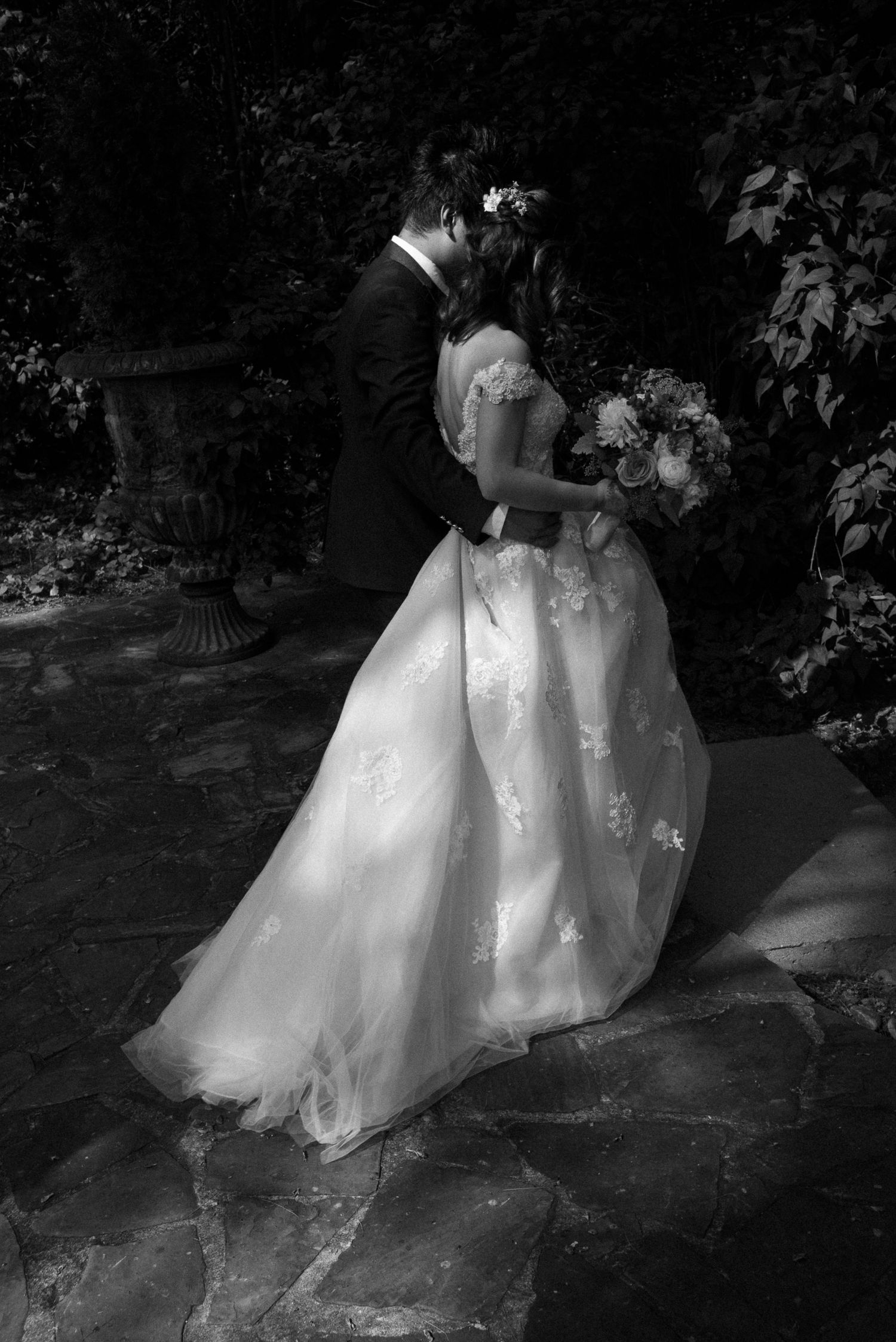 Black and white photo of a bride and groom sharing a kiss in a garden, bride holding a bouquet in a lace wedding gown.