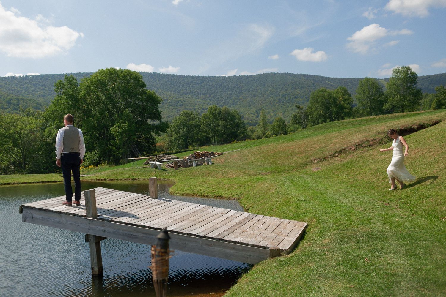 Man standing on wooden dock overlooking scenic mountain lake while woman in white dress walks on green hillside.