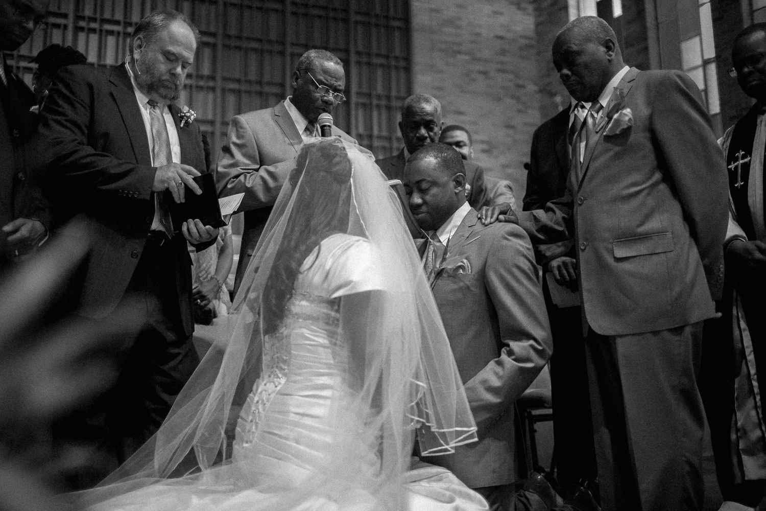 Black and white wedding photo of bride in veil surrounded by suited men praying on church steps.