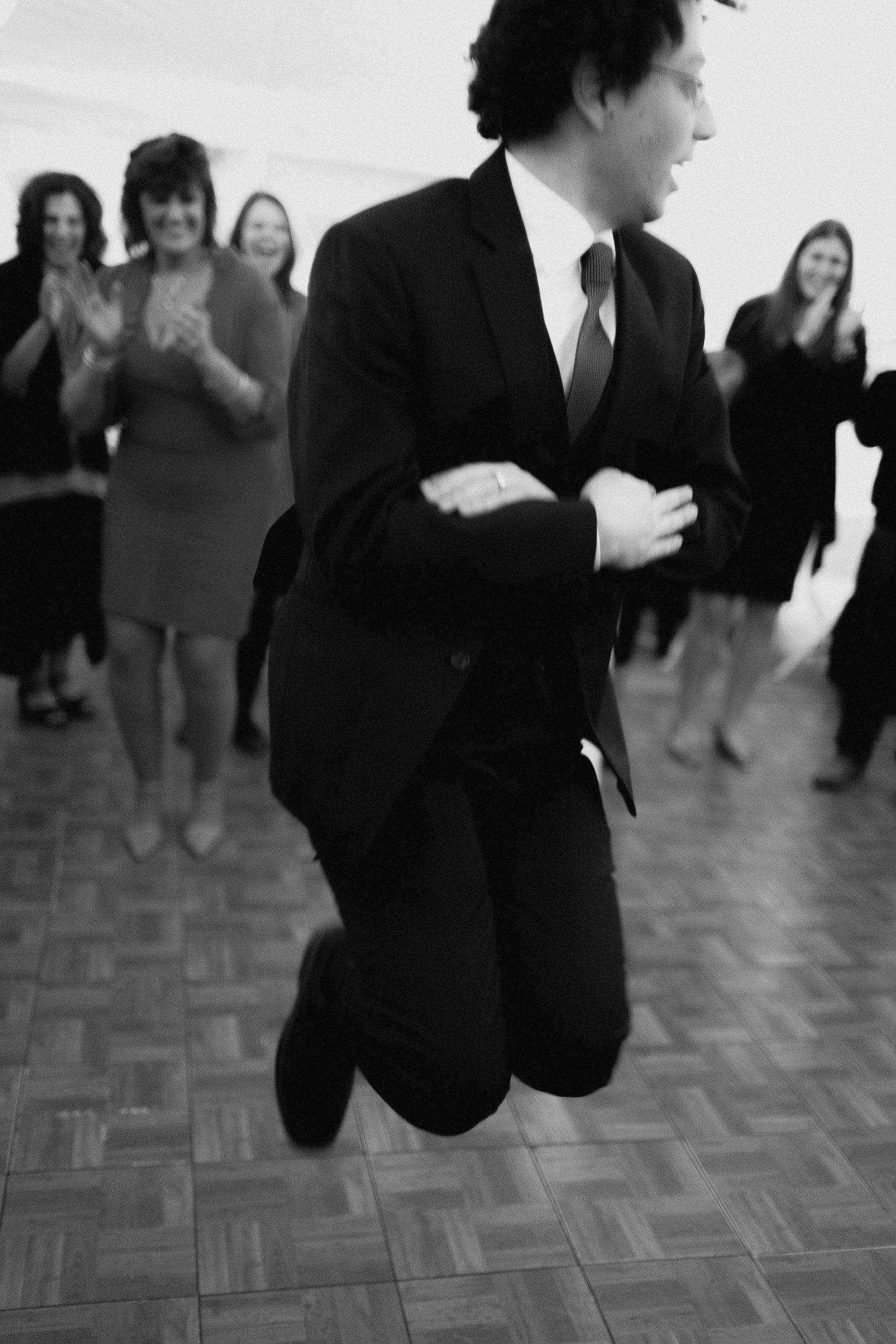 Man in suit jumping on dance floor while guests clap and smile in black and white photo.