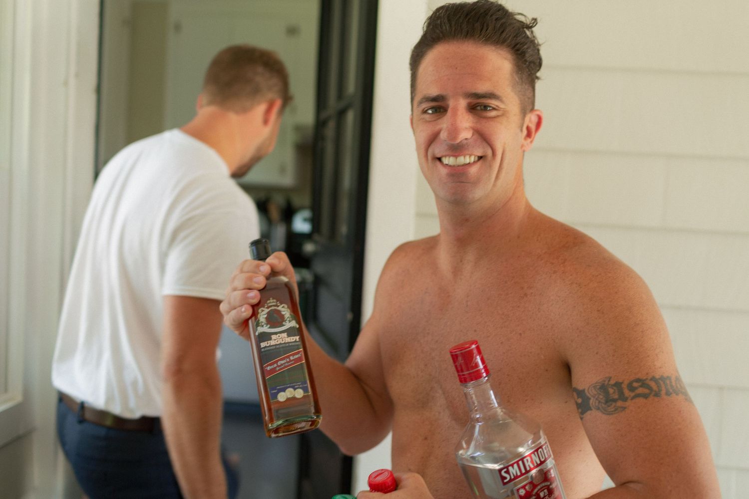 Shirtless man smiling while holding liquor bottles outdoors, with another man visible in the background.