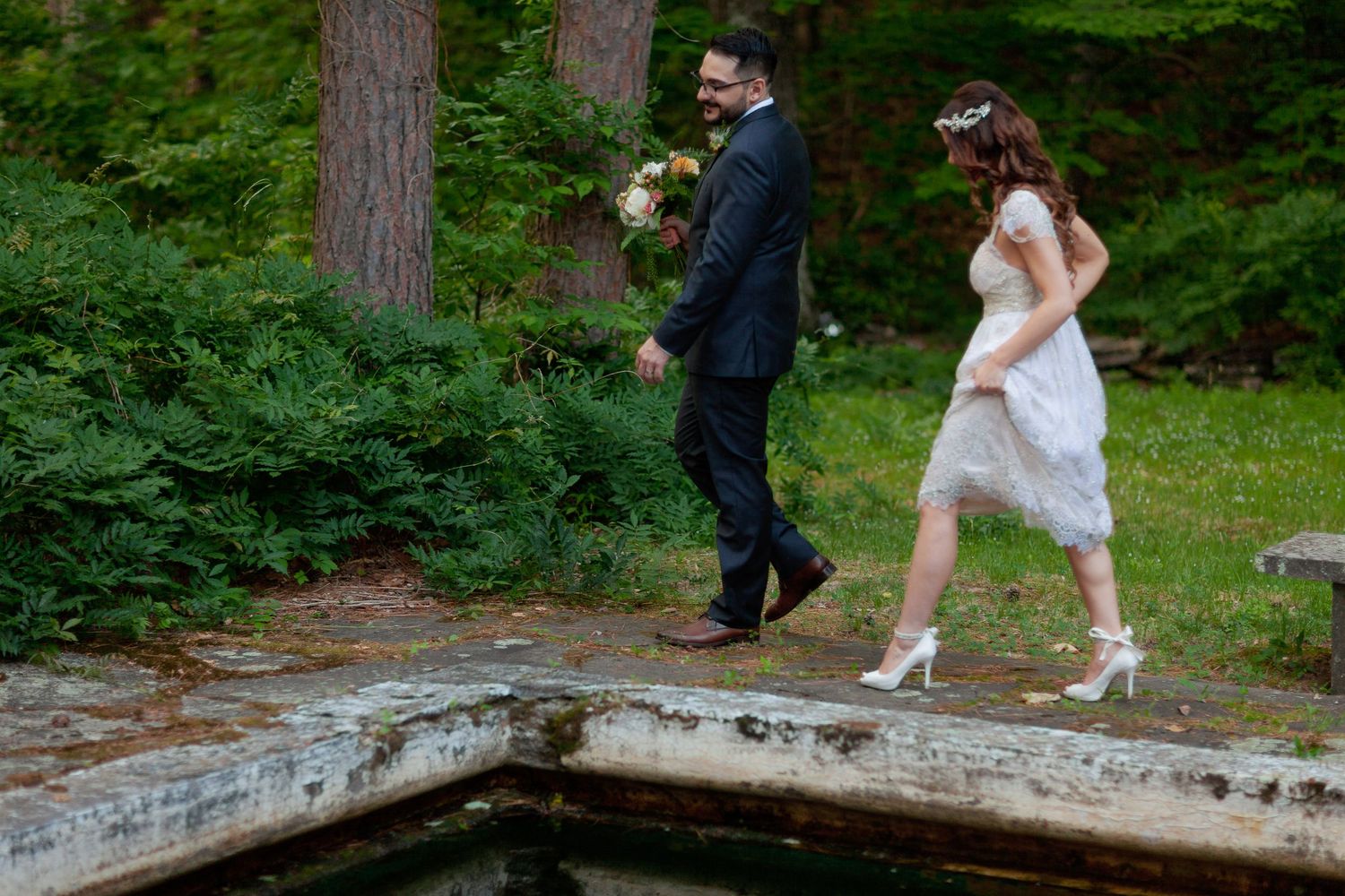 Bride and groom walking together outdoors near a pond in a lush green forest setting on their wedding day.