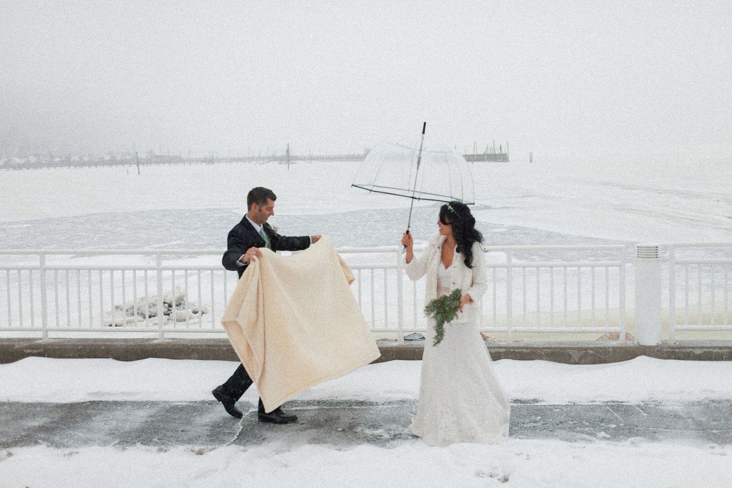 Bride and groom pose in snowy winter wedding photos outdoors by a frozen waterfront, bride holding a clear umbrella.