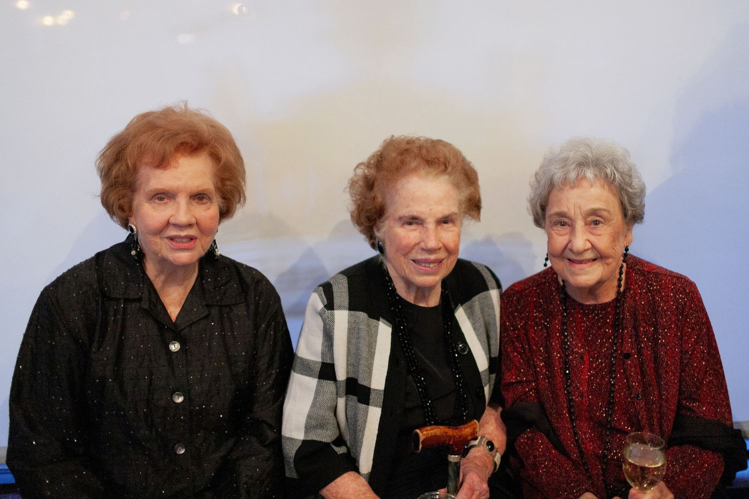 Three elderly women smiling together at a formal event, wearing elegant black and red outfits.