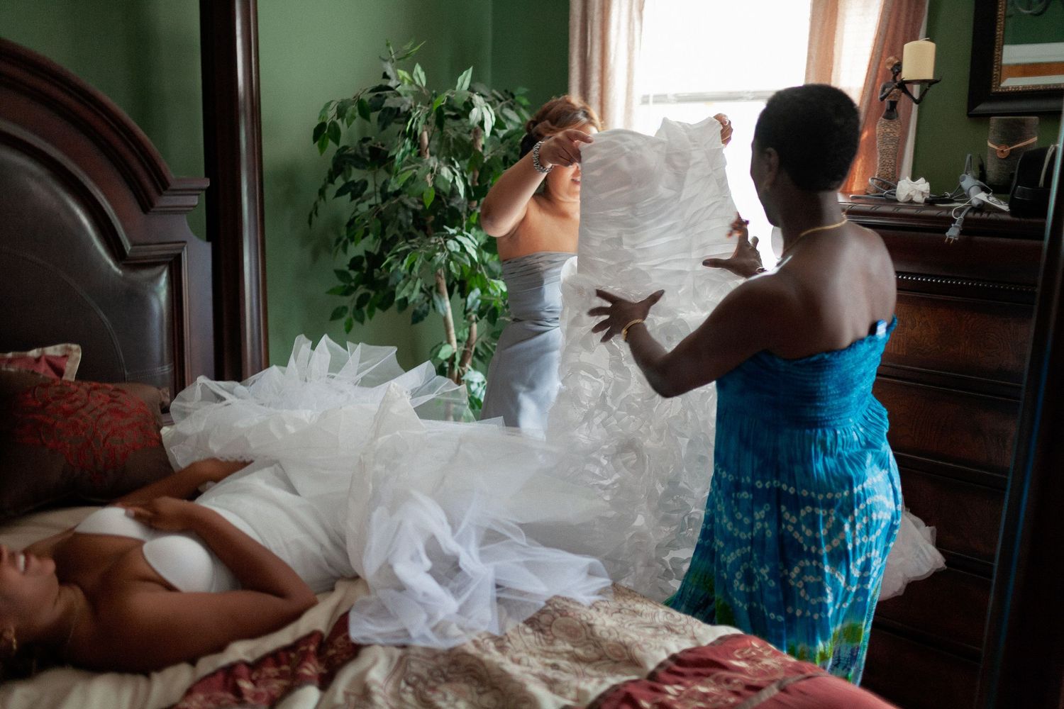 Bride getting help putting on white wedding dress in elegant bedroom with wooden furniture.