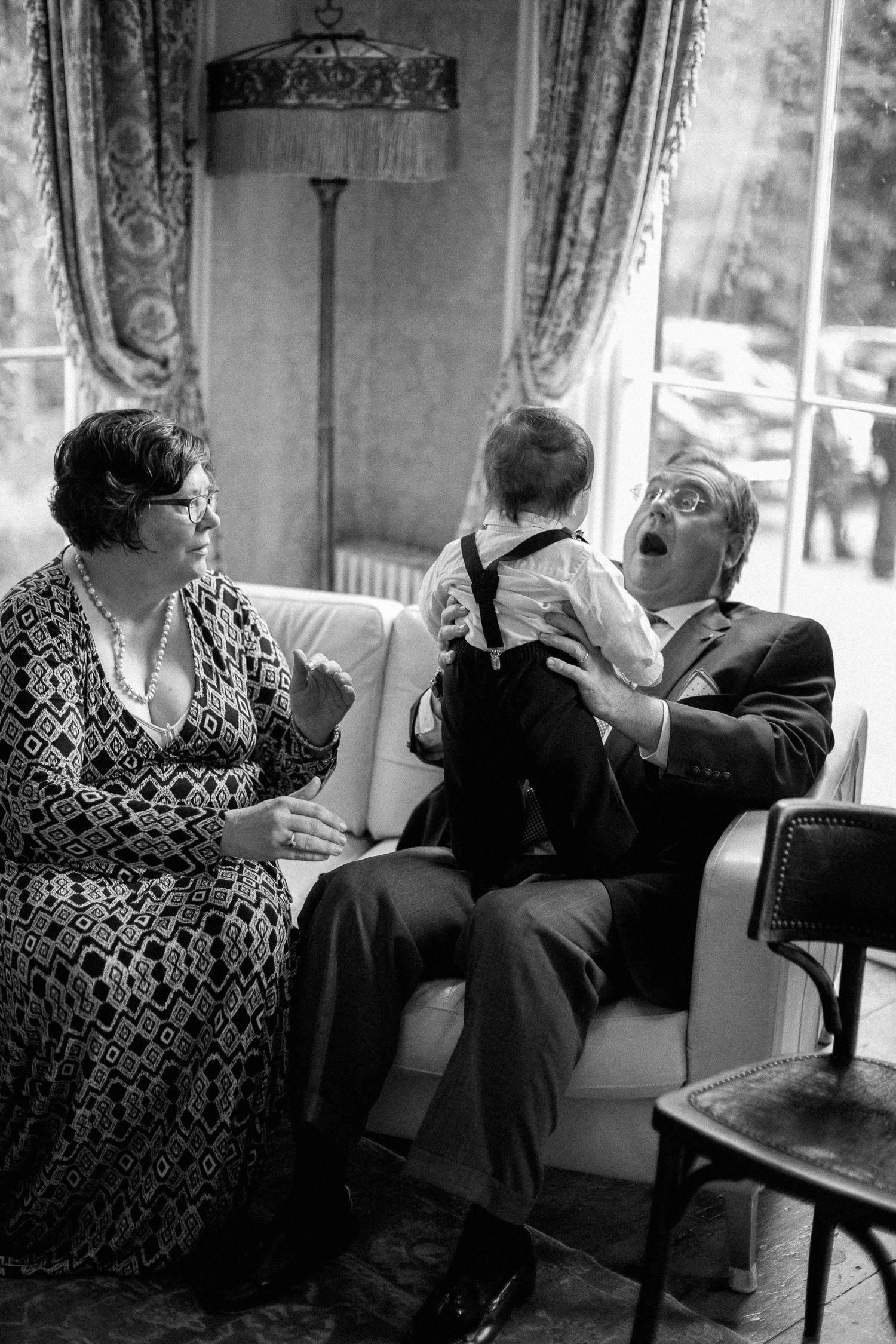 Black and white photo of grandparents sitting on sofa, joyfully interacting with a baby in suspenders.