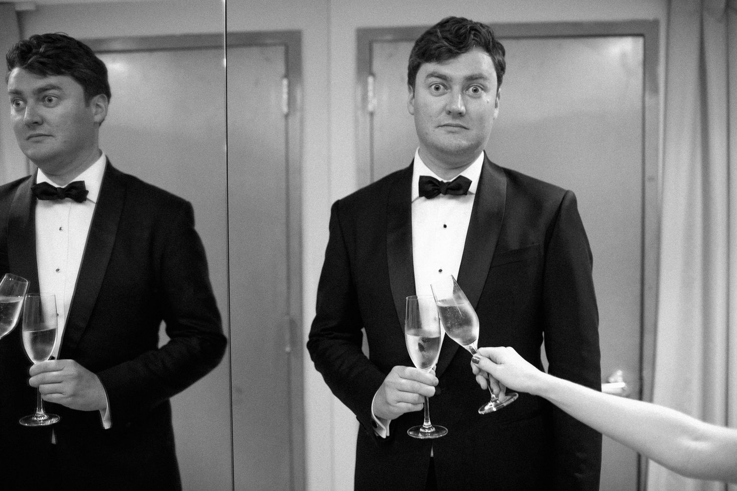 Man in tuxedo holding champagne glass at formal event, black and white photo with mirror reflection.