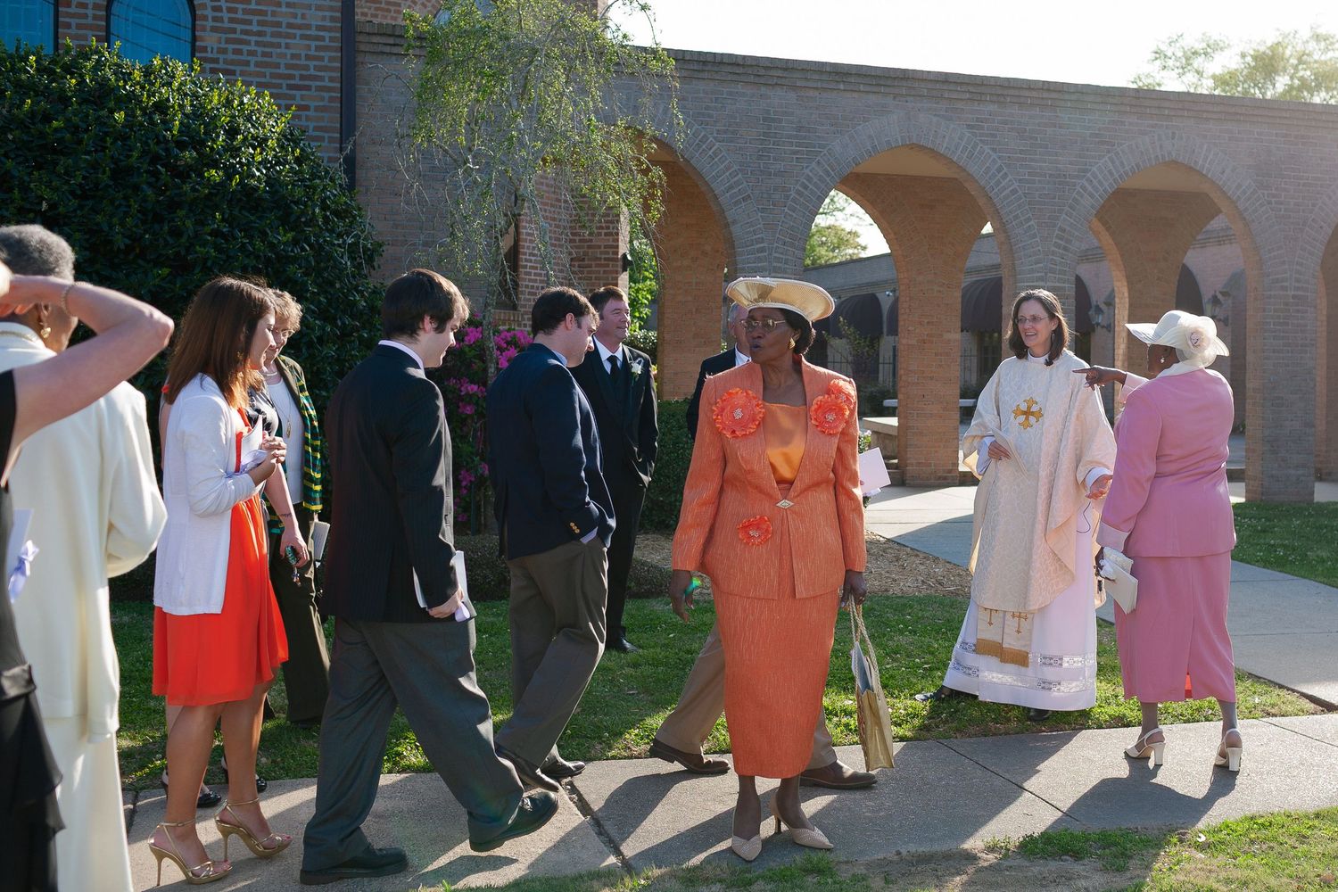 Group of people in formal attire walking outdoors near brick arches during a religious or ceremonial procession.