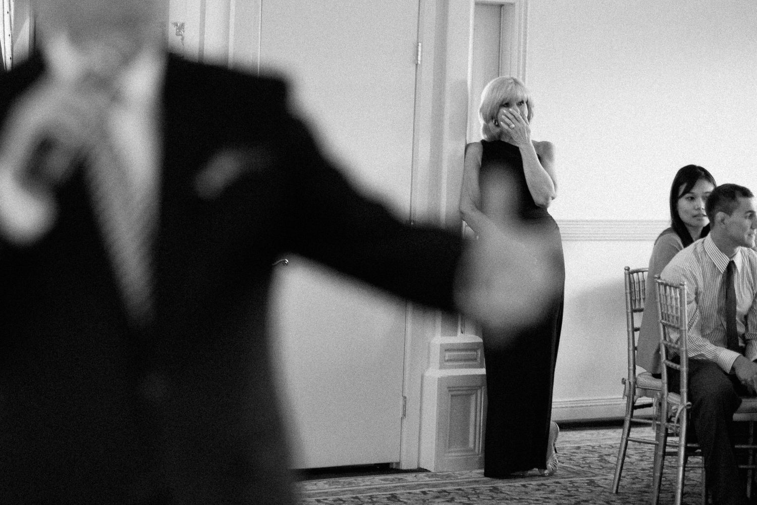 Black and white wedding photo of guests watching ceremony, woman in black dress covering mouth emotionally.