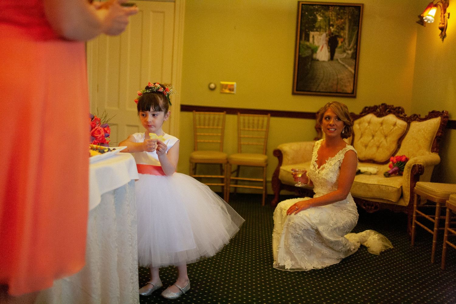 Bride in white gown kneels beside flower girl in tutu dress during wedding reception indoor setting.