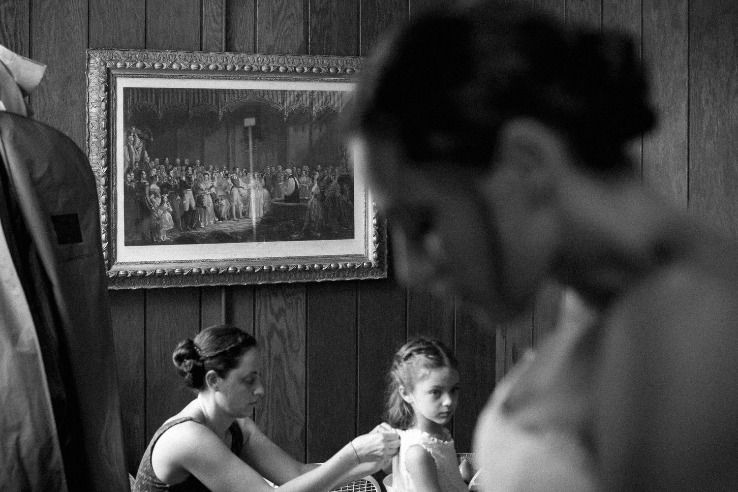 Black and white photo of women helping a young flower girl dress in a wood-paneled room with a framed painting on the wall.