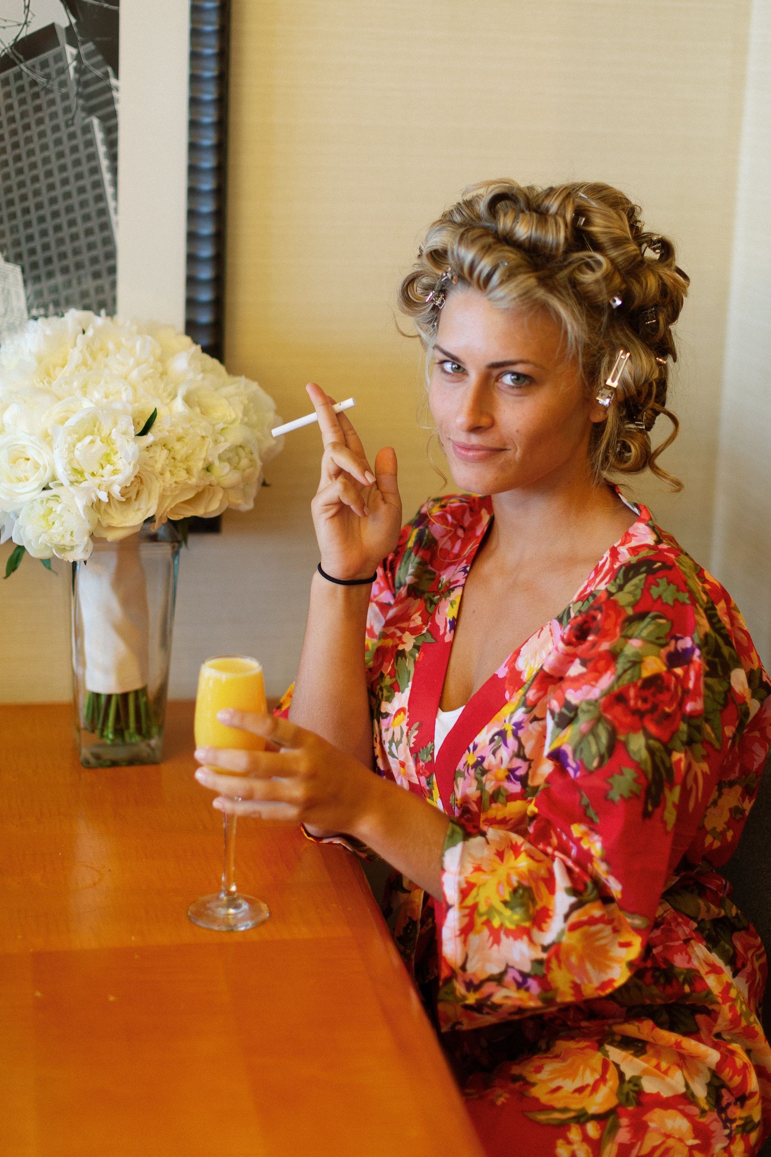 Bride in floral robe holding champagne and cigarette, with white roses bouquet, preparing for wedding day.