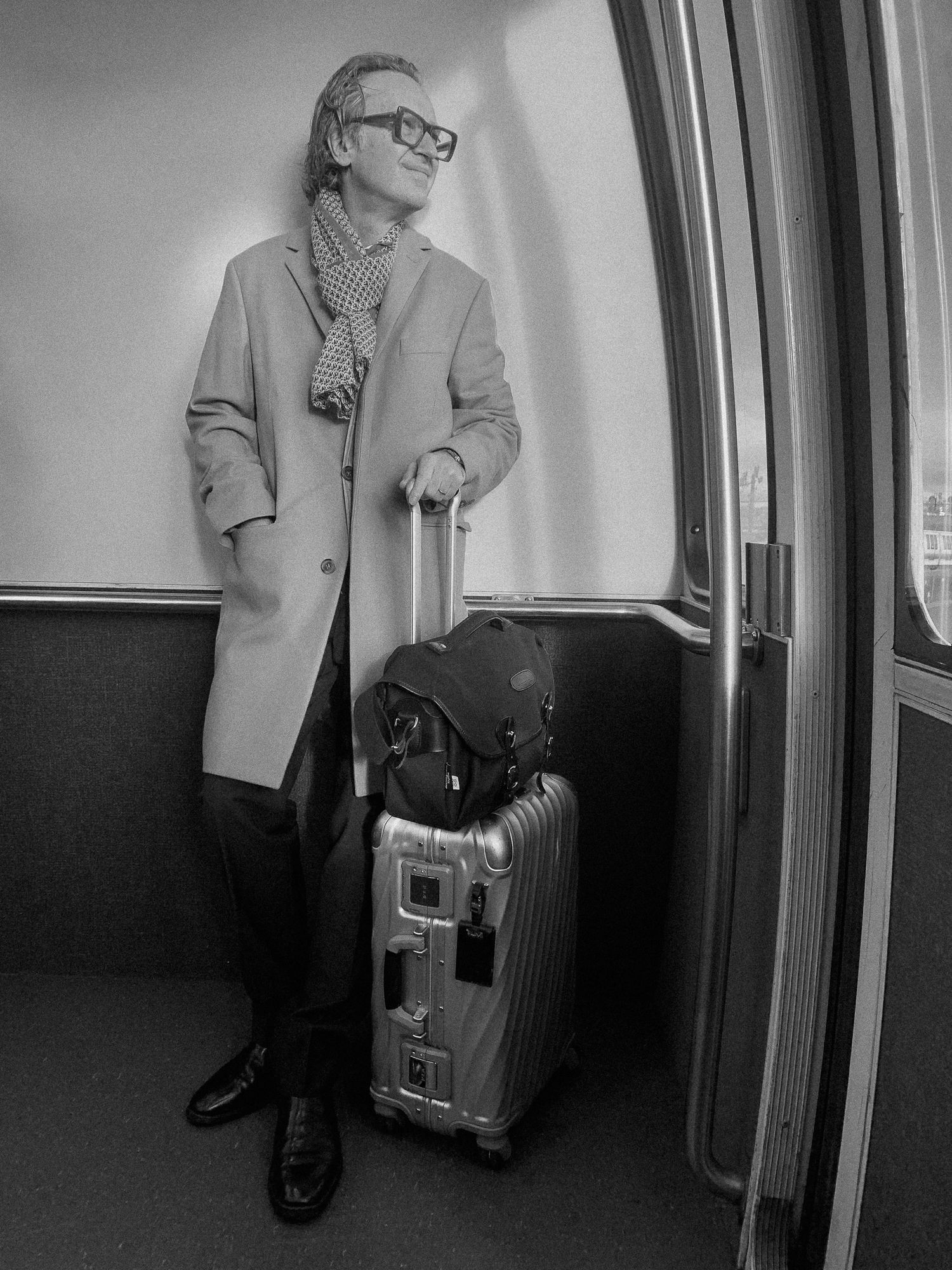 Man in long coat and scarf stands with Rimowa suitcase on train, black and white travel photography.