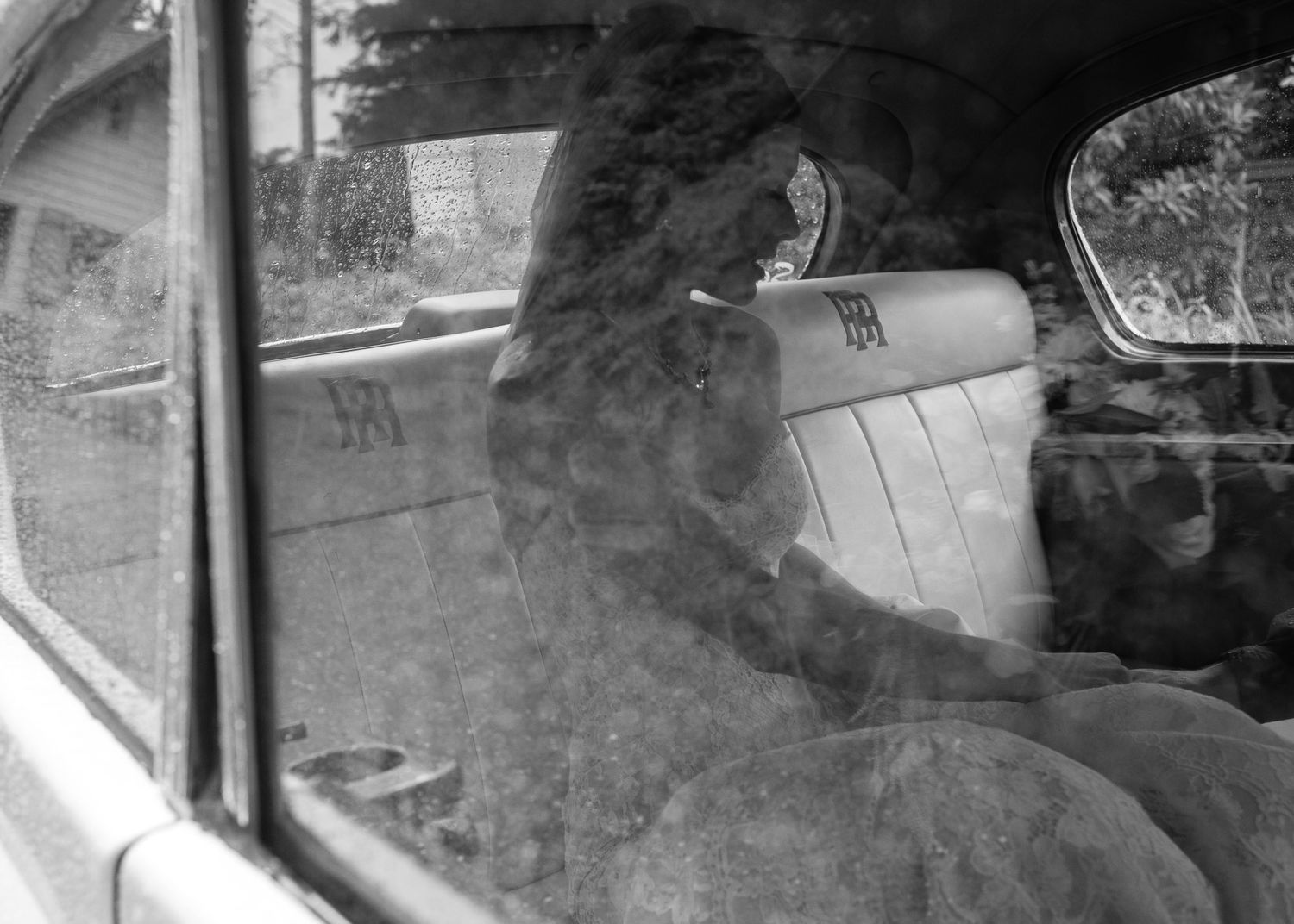A black and white photograph shows a person looking through a car window on a rainy day.
