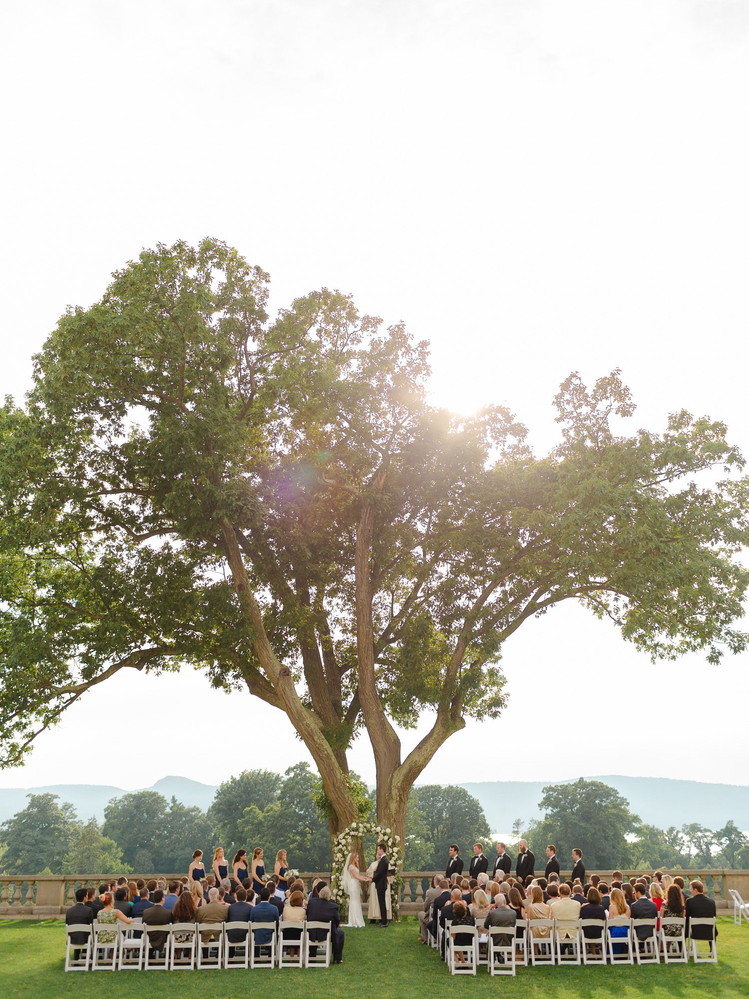 Panoramic view of an outdoor wedding ceremony setup under a large tree with guests seated on white chairs at sunset.