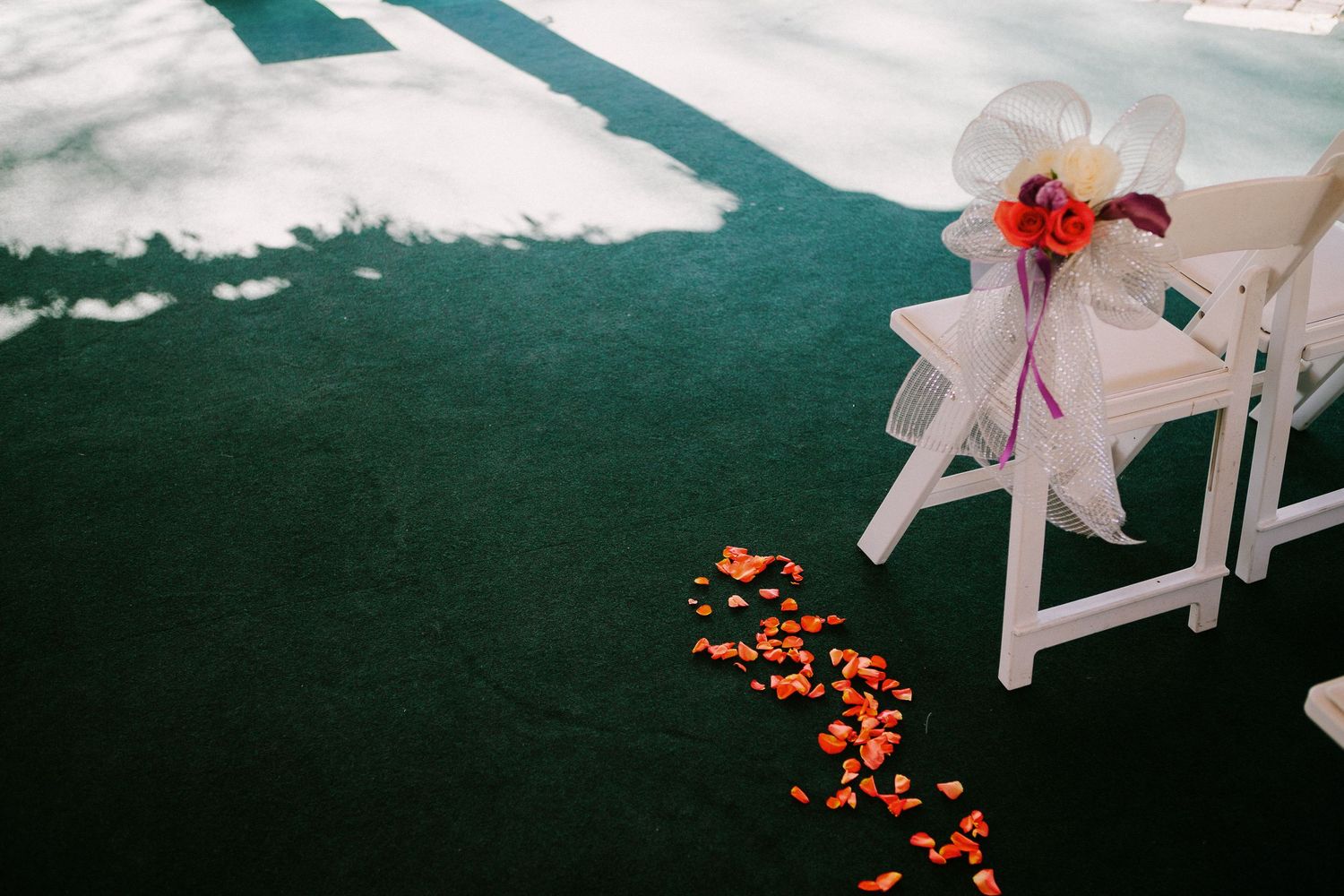 White folding chairs with floral decorations line a green carpet aisle scattered with orange and red rose petals.