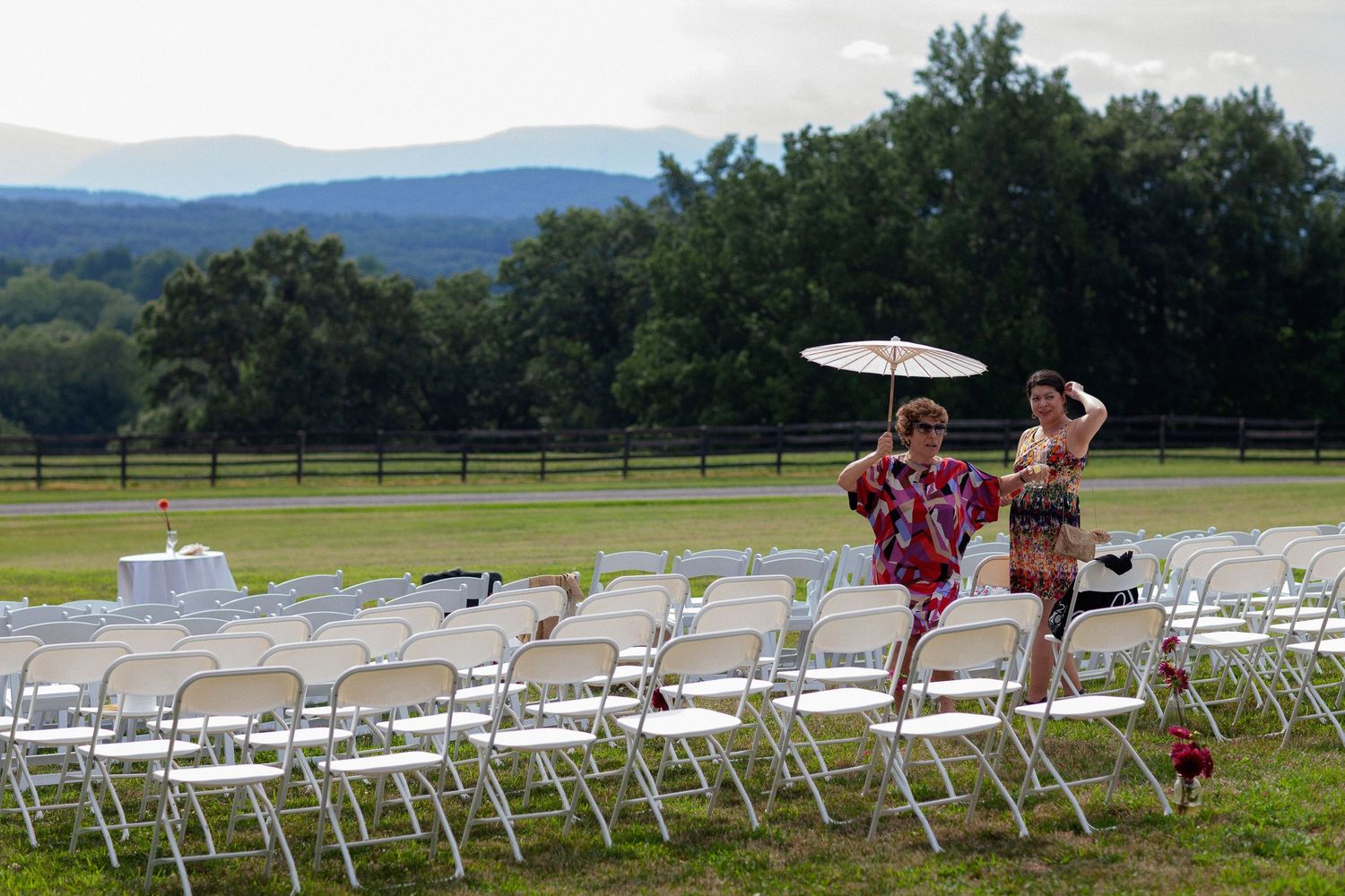 Outdoor wedding ceremony setup with white folding chairs on green lawn, mountains in background, two guests walking with umbrella.