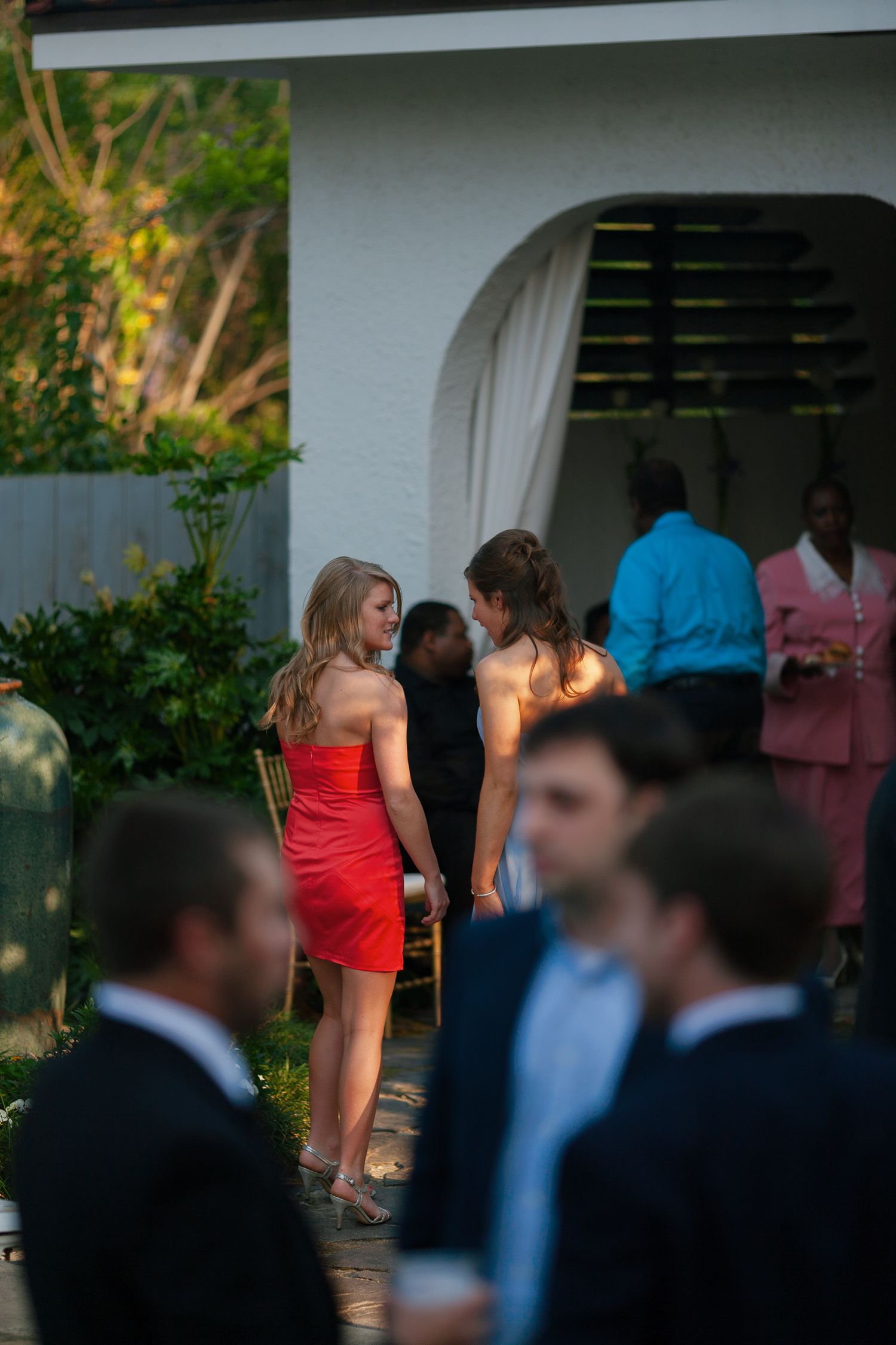 Two women in formal dresses chat at an outdoor evening event, with guests in suits visible in the foreground.