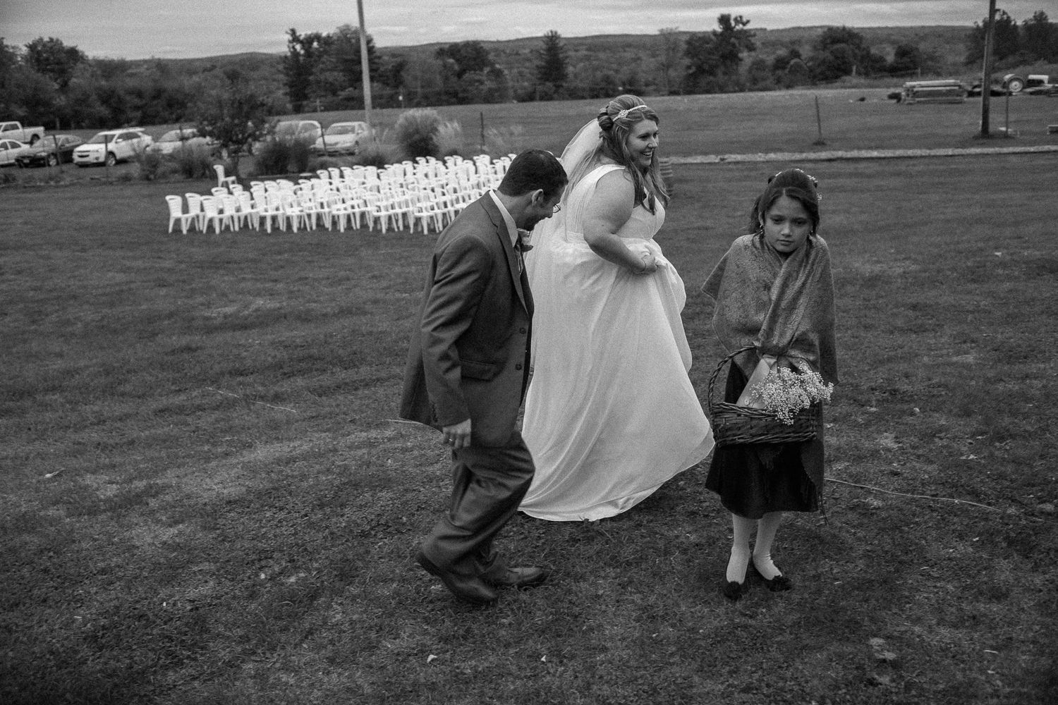 Black and white photo of a bride and groom walking with a young girl across a grass field at an outdoor wedding.