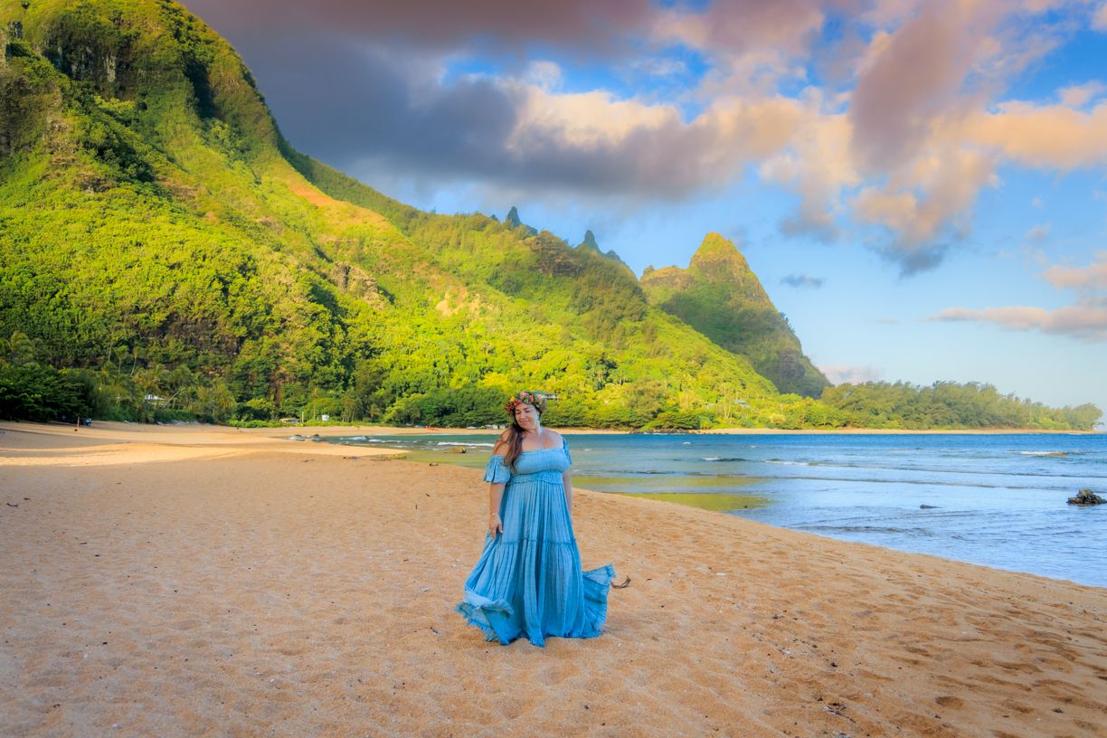 Woman in blue dress on sandy beach with lush green mountains and colorful sky in Hawaii.