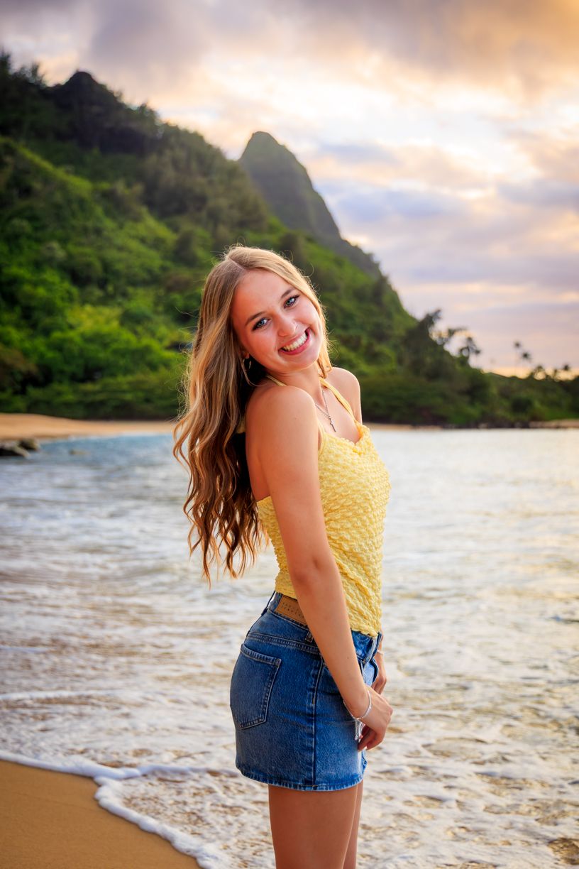 Woman in yellow top and denim shorts smiling on beach with ocean and mountains behind.