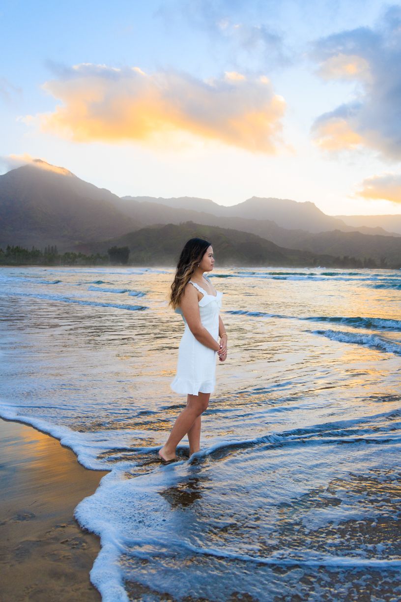 Woman in white dress standing in ocean waves at sunset with mountains in background.