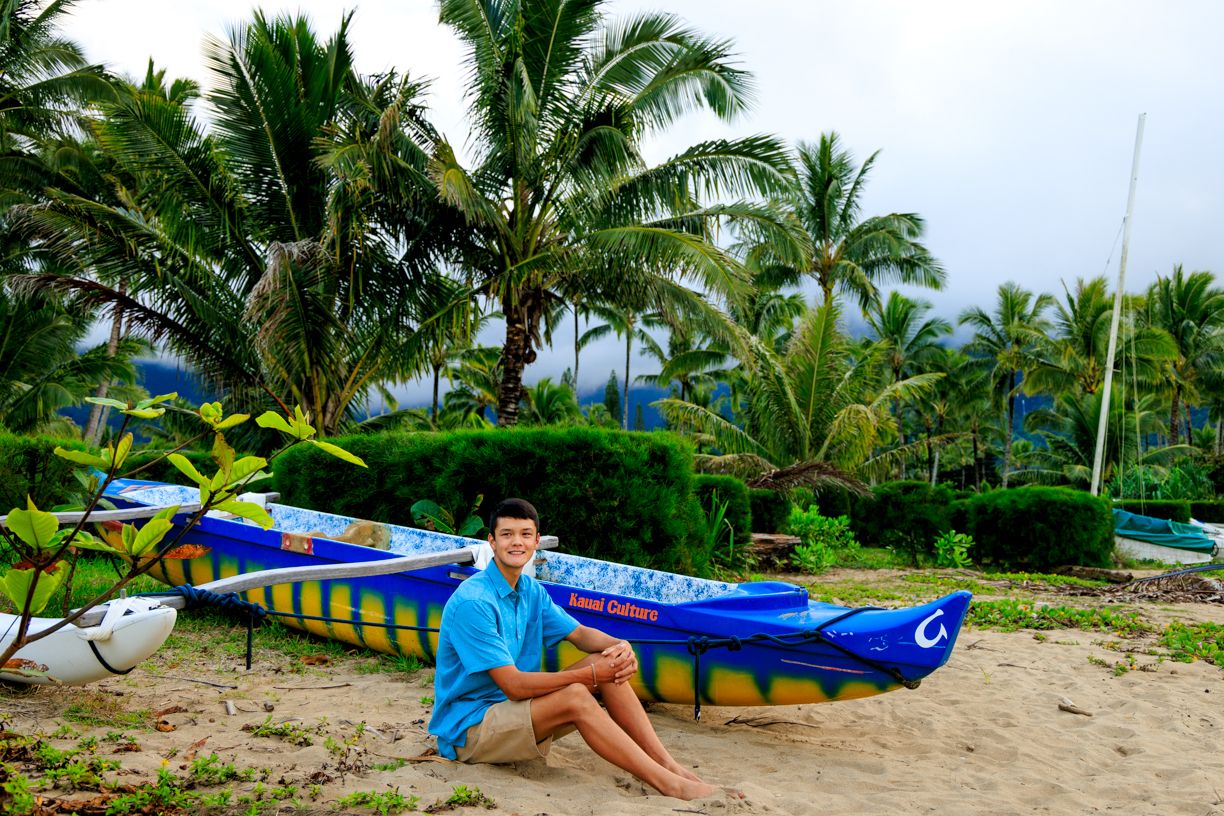 Man in blue outfit sitting on blue kayak on beach with palm trees in background.