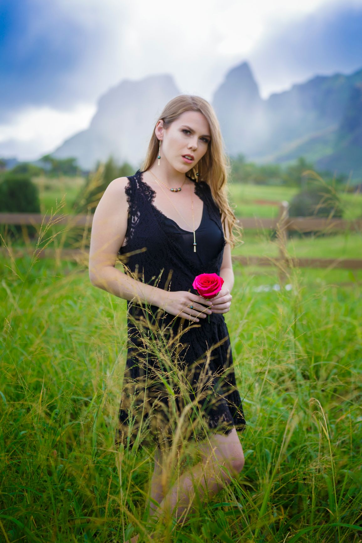 Woman in black dress holding red rose poses in field with misty mountains in background.