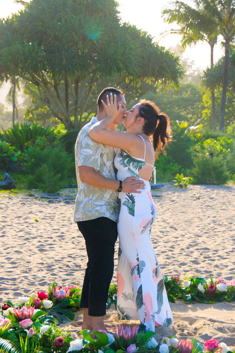 A romantic beach proposal scene with pink flowers arranged in a circle on the sand at sunset.