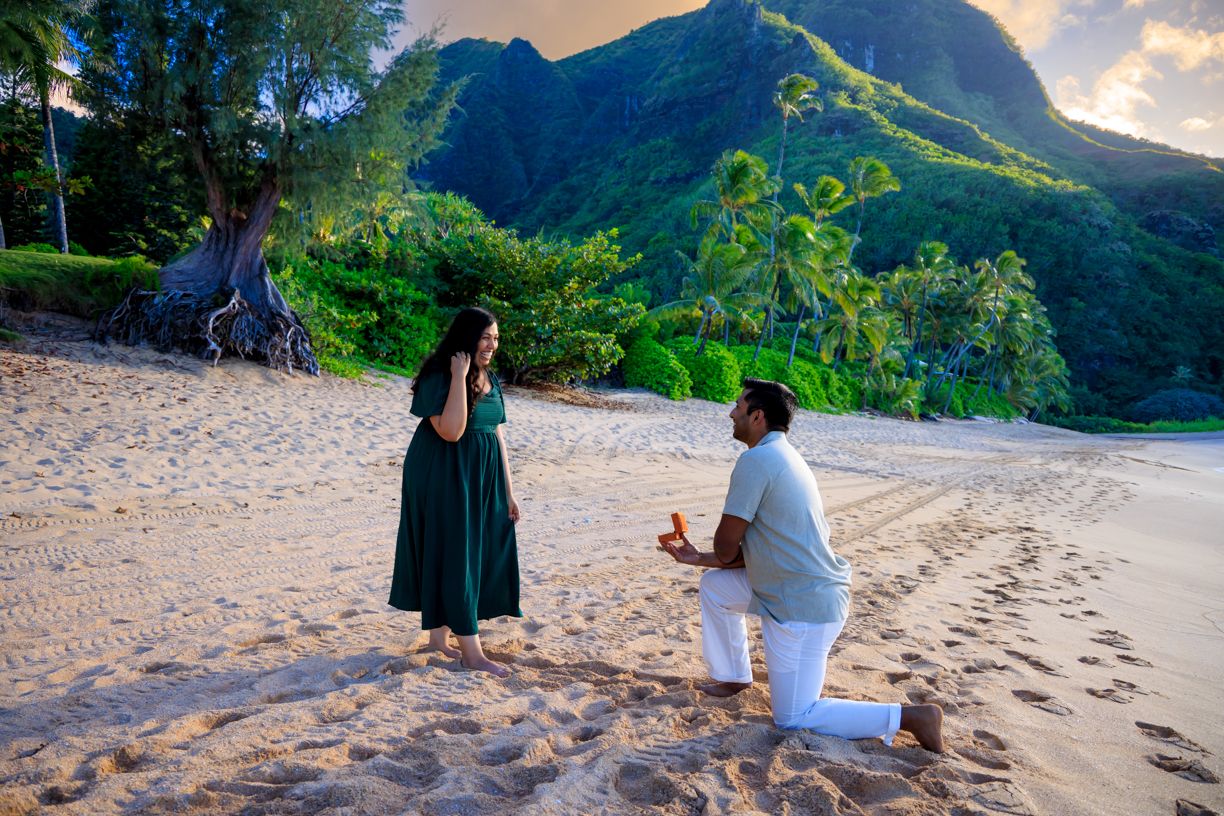 A couple shares romantic moments on a tropical beach with mountains and palm trees in the background.